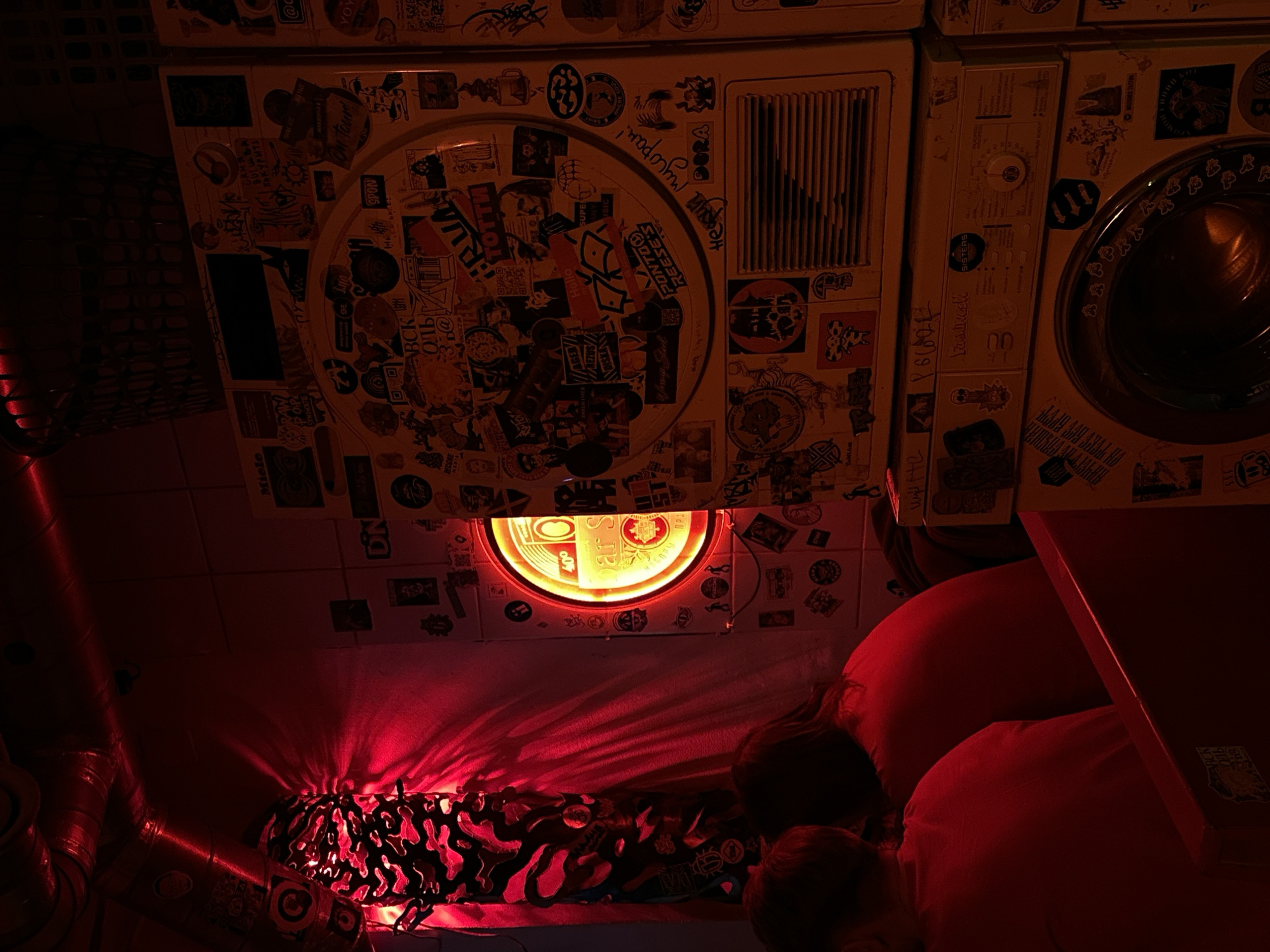 The image shows a dimly lit scene in what appears to be an interior space, possibly a bar or restaurant. The primary light source casts a red hue throughout the area, emanating from below the ceiling. The ceiling is covered with two washing machines covered in stickers. Below the washing machines is a round light fixture embedded in the ceiling, radiating a warm orange color. Streaks of red light flood the area below, illuminating seating in the form of red cushions or pillows, where two heads of people are dimly visible. A corner of a red table is partially visible in the right foreground. A red pipe runs diagonally along the left side of the frame, and a patterned lamp or light fixture lies underneath the light fixture on the ceiling. The washing machines have stickers from what appears to be the Russian Federation, and is likely located in the city of Saint Petersburg, Russia.