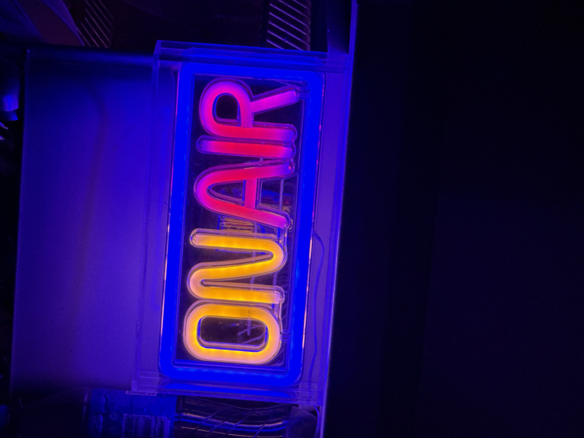 An "ON AIR" neon sign is illuminated with yellow and pink lights, encased in a clear plastic box with a blue neon border. The sign is positioned in a dimly lit environment, likely indoors, with a dark background. The upper portion of the image shows a purple-hued surface, possibly a wall or ceiling. The sign itself is the primary subject, with the bright neon lights contrasting against the darker surroundings. The image does not contain any information about the city of Drianovets, Bulgaria, or any people.