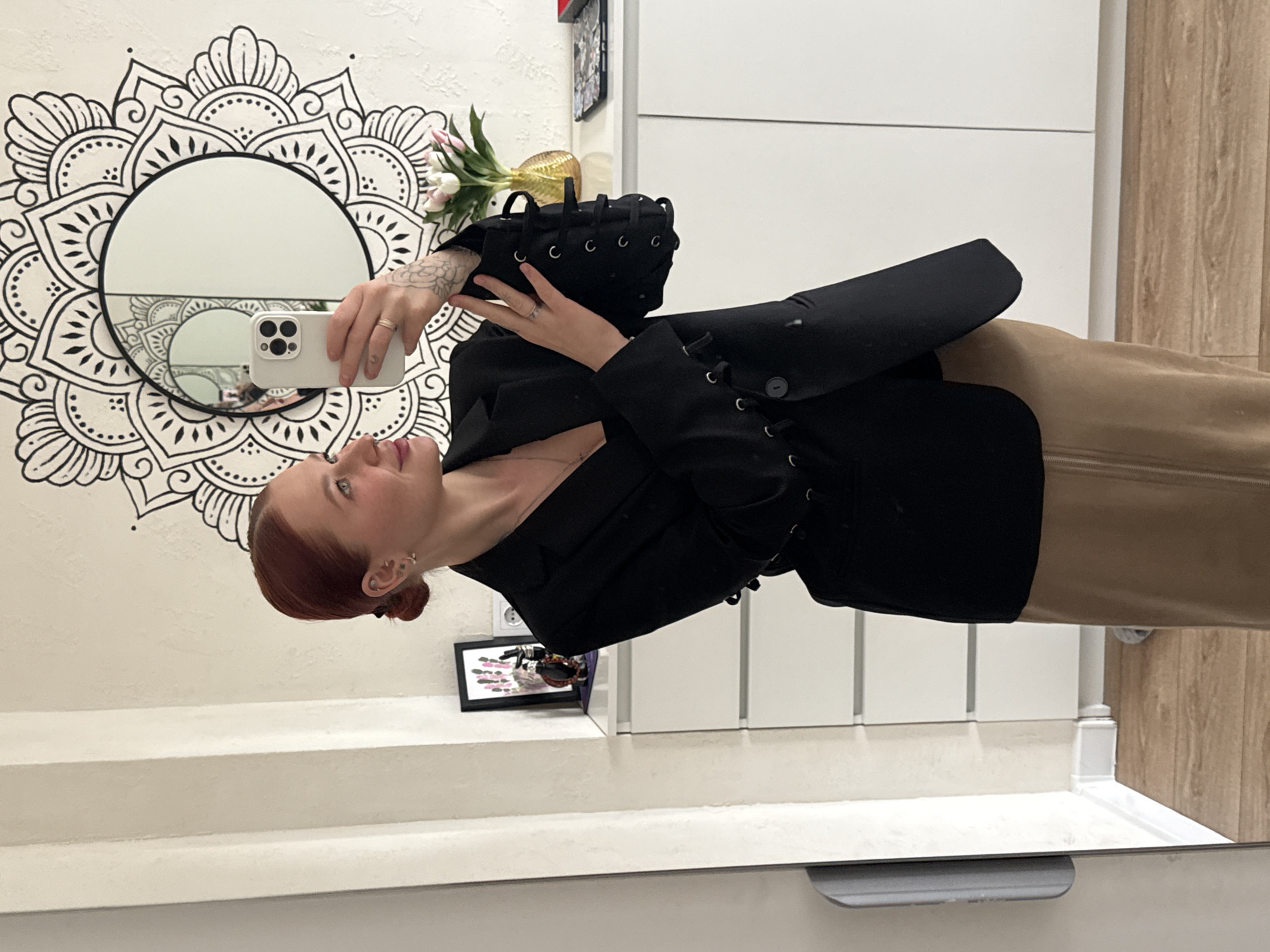 A woman with auburn hair is taking a mirror selfie with a white smartphone, wearing a black blazer-style top with lace-up detailing and a tan skirt or pants. The mirror hangs on a white wall decorated with a black and white floral mandala design. To the right of the woman is a white cabinet or wardrobe with vertical seams. A vase with flowers rests on top of the cabinet. Below the mirror, there is a white shelf with a framed photo or artwork. The location is presumed to be an interior space in Rostov-on-Don, Russia. The image includes a portion of the mirror's edge at the bottom, suggesting it is a reflection of the bathroom.