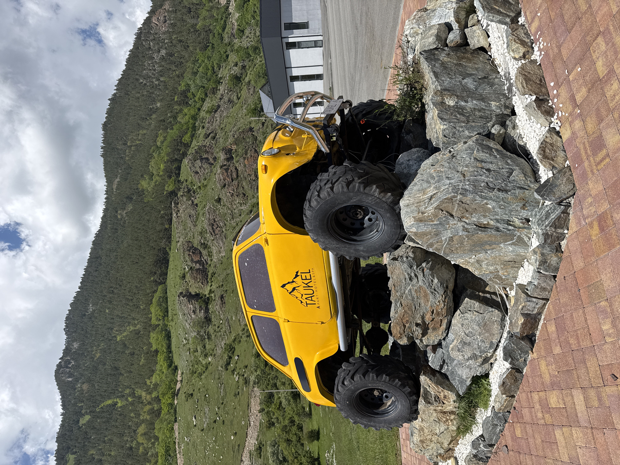 In Teberda, Russia, a bright yellow, modified car with oversized tires is prominently displayed on a rocky mound, serving as a static exhibit. The vehicle, a classic model, has been significantly altered with a lifted chassis and rugged off-road tires, suggesting it is no longer functional for regular use. It is adorned with a logo on its side that reads "TAUKEL APART-HOTEL & SPA," indicating its connection to a local establishment.
The car is positioned on a carefully arranged collection of large, grey stones, which are themselves surrounded by white gravel and a border of smaller stones. This landscaping is set against a backdrop of a paved area, likely a parking lot, and a modern building with a dark roof and white walls featuring multiple windows.
Beyond the immediate display, the environment transitions into a lush, green landscape. Rolling hills covered with dense pine forests rise in the background, with patches of lighter green foliage and rocky outcrops visible on their slopes. The sky above is a mix of white clouds and patches of blue, suggesting a partly cloudy day. The overall scene appears to be an outdoor area intended for visual appeal and to attract visitors, possibly associated with the hotel. No people are visible in the frame.