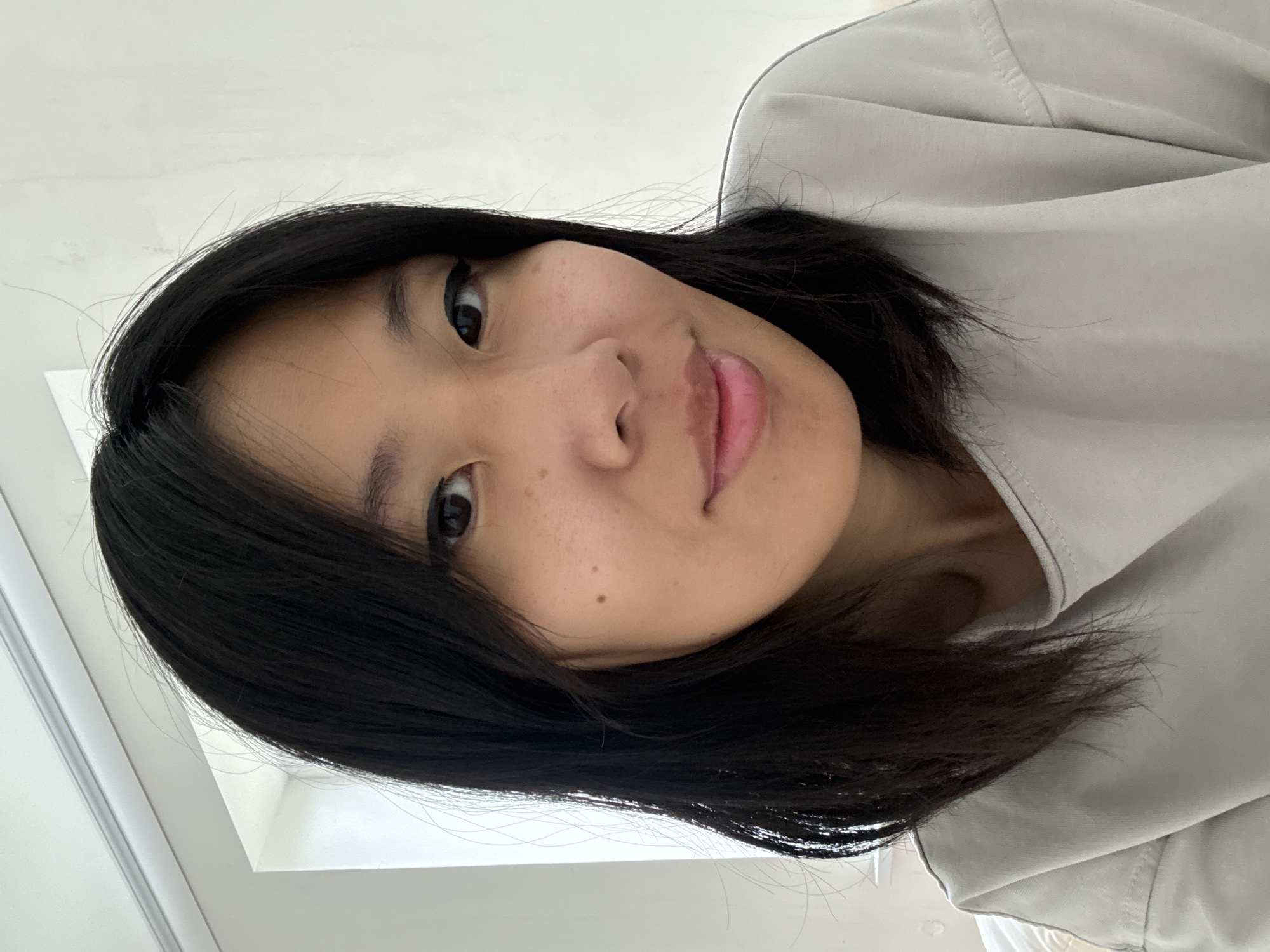 A close-up, slightly rotated selfie of a woman with dark, shoulder-length hair. She has fair skin with a few freckles. Her eyes are dark, and she is wearing subtle eyeliner. Her lips are slightly parted. She is wearing a light gray shirt or top. The background appears to be a white wall, possibly indoors. The location is not discernible from the image. Ембаево, Russia cannot be determined from the image.