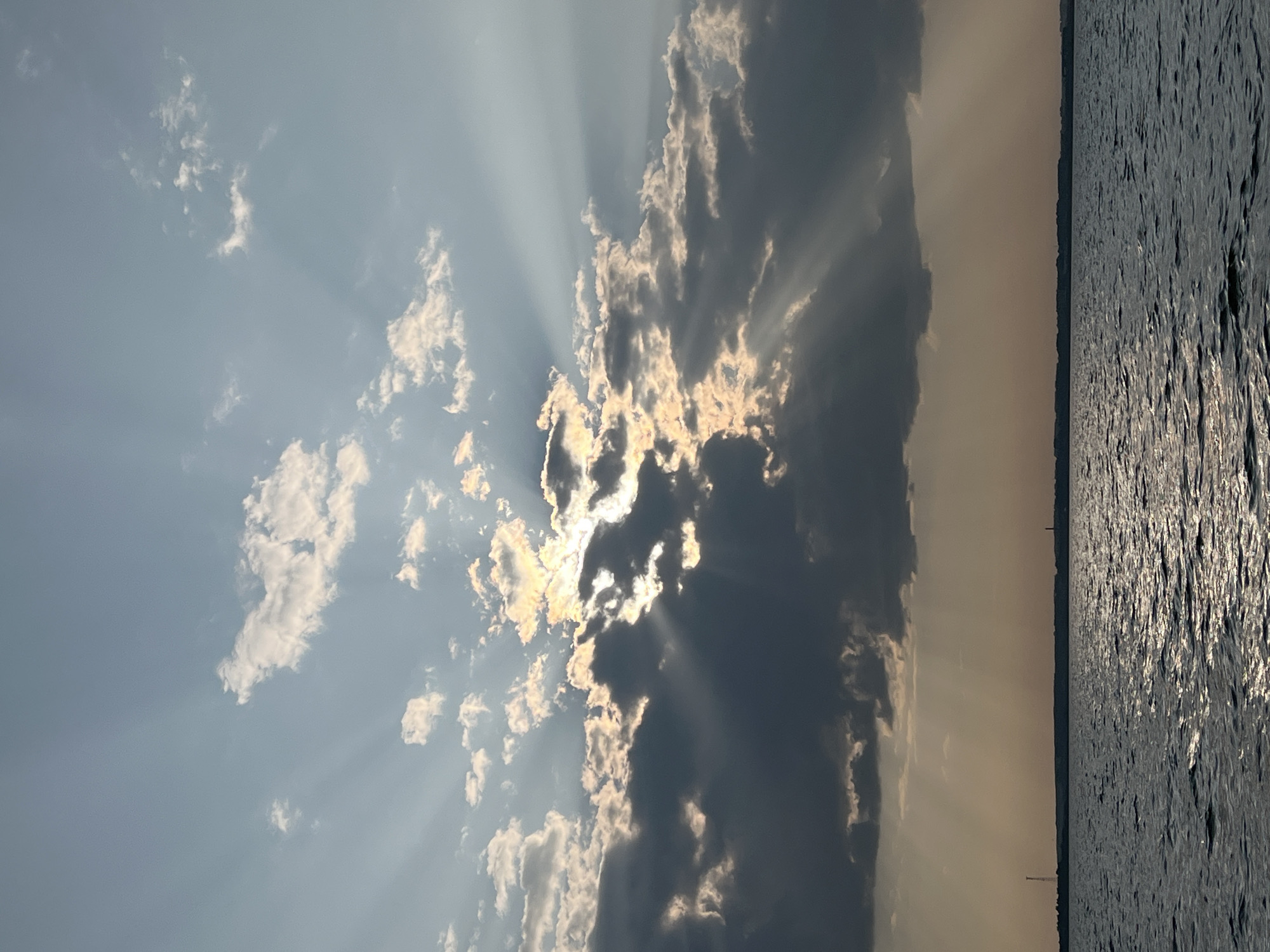 The image depicts a wide-angle view of a body of water under a dramatic sky. Dark, heavy clouds dominate the middle portion of the sky, with shafts of sunlight piercing through them, creating a crepuscular ray effect. Lighter clouds are scattered above and around the darker ones. The sun itself is partially obscured by clouds. The horizon line, visible in the lower third of the image, is a distant, dark silhouette of land, possibly with faint structures such as towers or buildings. The water in the foreground is choppy, reflecting the muted sunlight and creating a textured surface. The overall impression is one of a powerful, natural light display over a serene, expansive landscape, characteristic of a sunset or sunrise over a large body of water. The specific location in Novopichugovo, Russia, implies a natural setting such as a lake or a wide river.