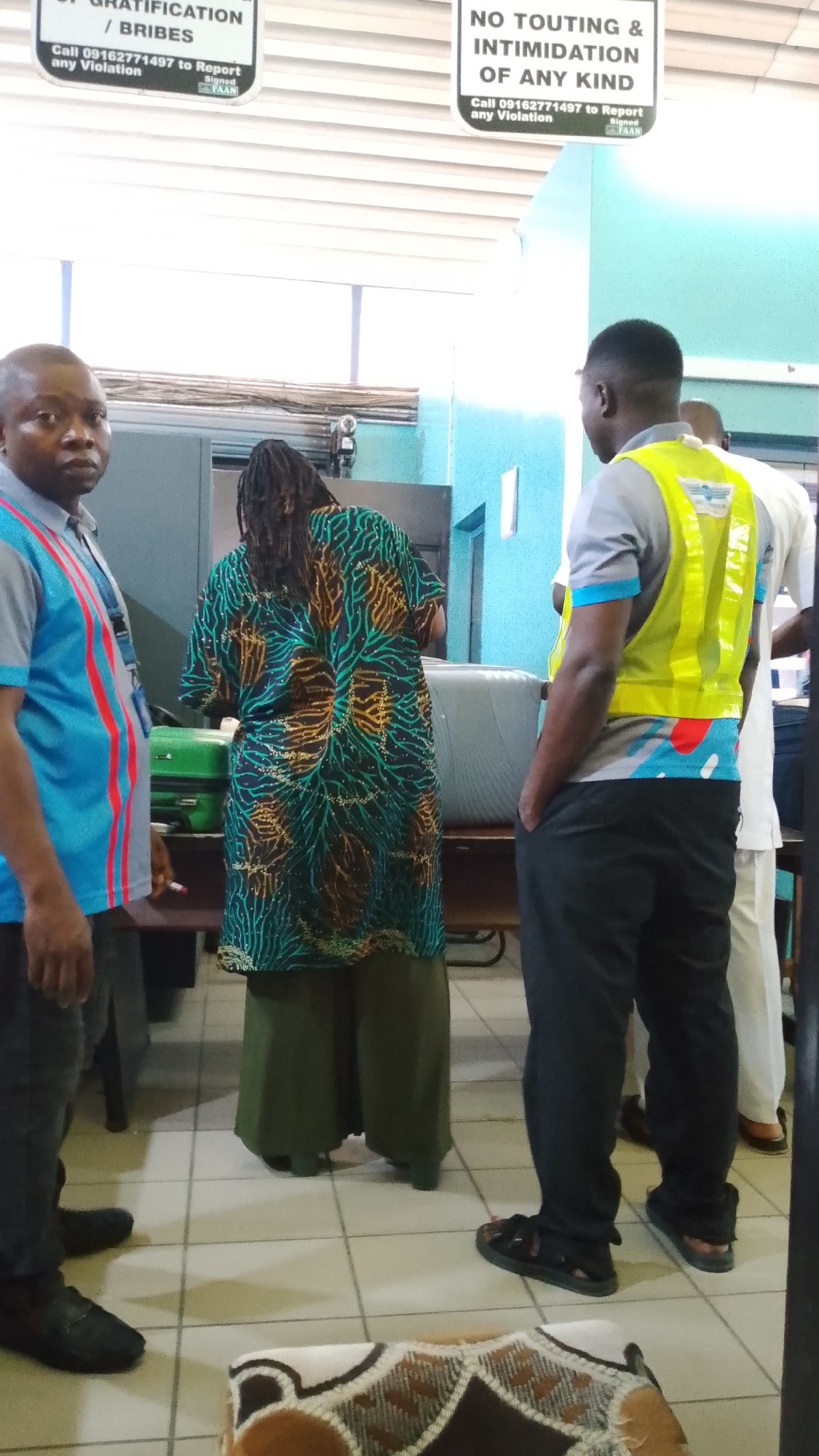 The image depicts an indoor scene, likely a public facility in Orerokpe, Nigeria. In the foreground, a man with dark skin and a shaved head, wearing a blue and red striped shirt, is partially visible on the left. To his right, a person with dreadlocks is seen from behind, wearing a vibrantly patterned green, teal, and orange tunic over olive-green wide-leg pants. In the center-right, a man with dark skin, wearing a grey t-shirt with a blue collar and a bright yellow safety vest, stands with his hands in his pockets, facing away from the camera. Behind him, another person in white attire is partially visible. The background shows a tiled floor, a dark counter or table, and metal shelving units. Two pieces of luggage, one green and one grey, are placed on the counter. Two signs are visible on the wall above the counter, one in black and white with text including "OF GRATIFICATION / BRIBERIES" and "Call 09162771497 to Report any Violation," and another sign with text that includes "NO TOUTING & INTIMIDATION OF ANY KIND." The overall impression is a waiting area or service point.