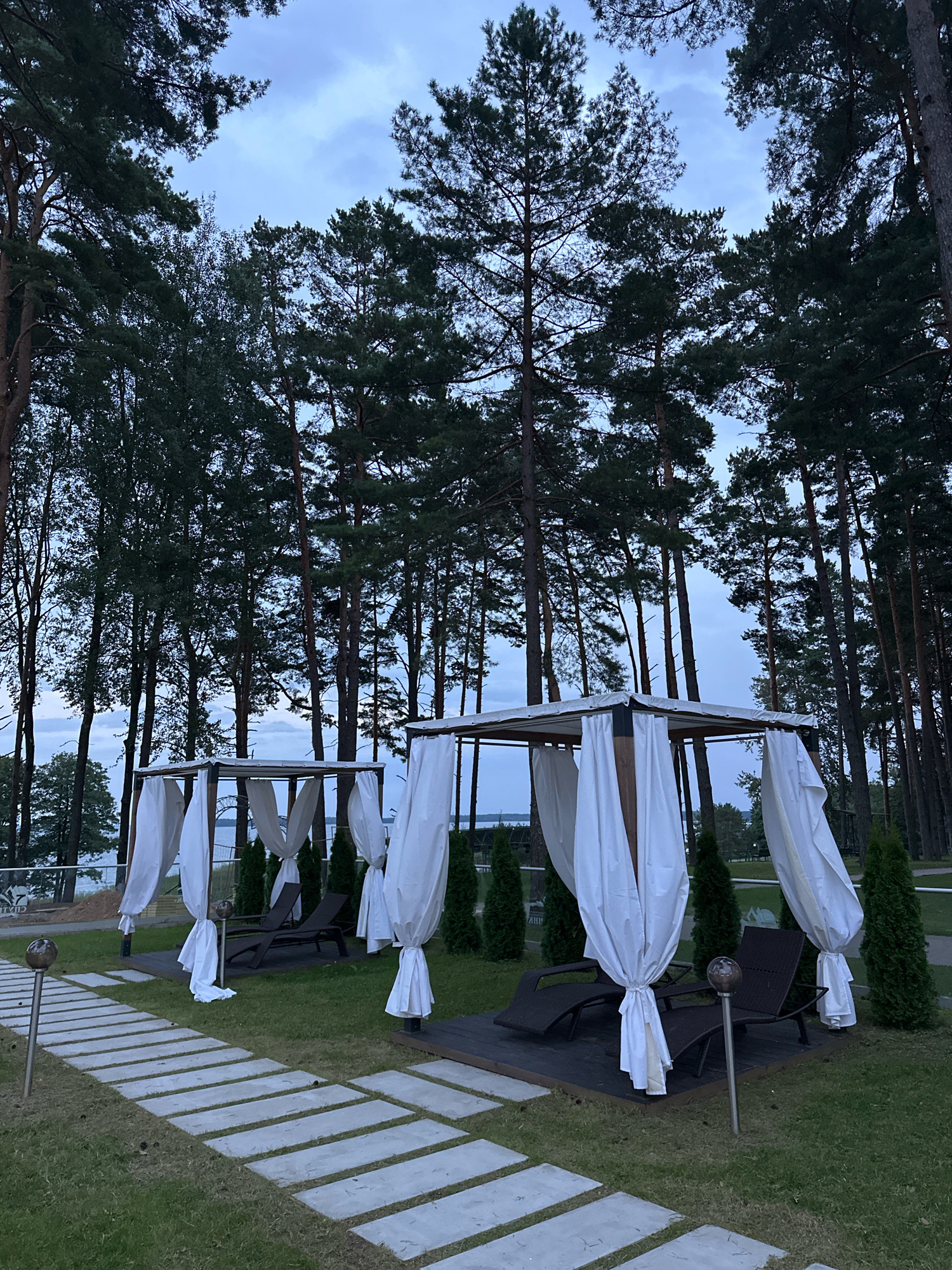 The image depicts an outdoor seating area situated in a forested location. Two open-sided gazebos, each draped with white fabric curtains, are prominently featured. Each gazebo contains two dark-colored lounge chairs. A stone pathway runs through the grassy area in front of the gazebos. In the background, tall pine trees dominate the landscape, with a body of water and a hazy sky visible beyond them. The overall atmosphere suggests a tranquil, possibly resort-like, environment, likely in the city of Нарацкі сельскі Савет, Belarus, given the visual cues of pine forests and a large body of water. There are no people visible in the image.