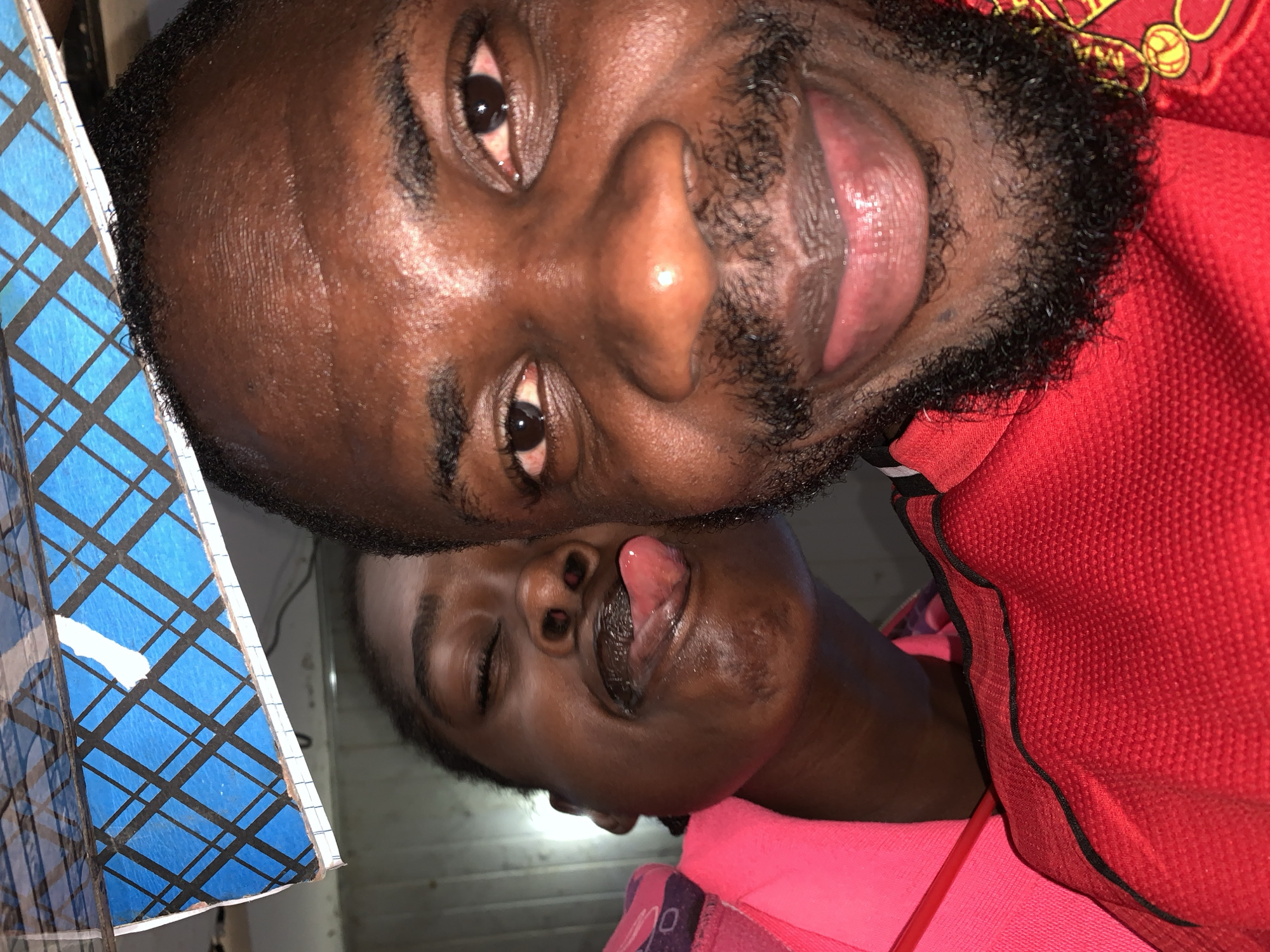 This is a close-up, overhead shot of two Black males in what appears to be a bedroom in New Stadium, Nigeria. The lighting is dim, suggesting an indoor setting. The male in the foreground, to the left of the image, is positioned looking up, with his mouth slightly open and his tongue extended. He has a beard and dark, curly hair. He is wearing a red shirt with black trim. The male behind him, to the right, is also looking up and appears to be asleep or unconscious, with his eyes closed and his mouth slightly agape. He is wearing a bright pink garment. A blue and black checkered notebook is visible to the far right of the image, partially obscuring the background. The overall impression is one of intimacy or possibly distress, given the unusual positioning and expressions of the individuals.