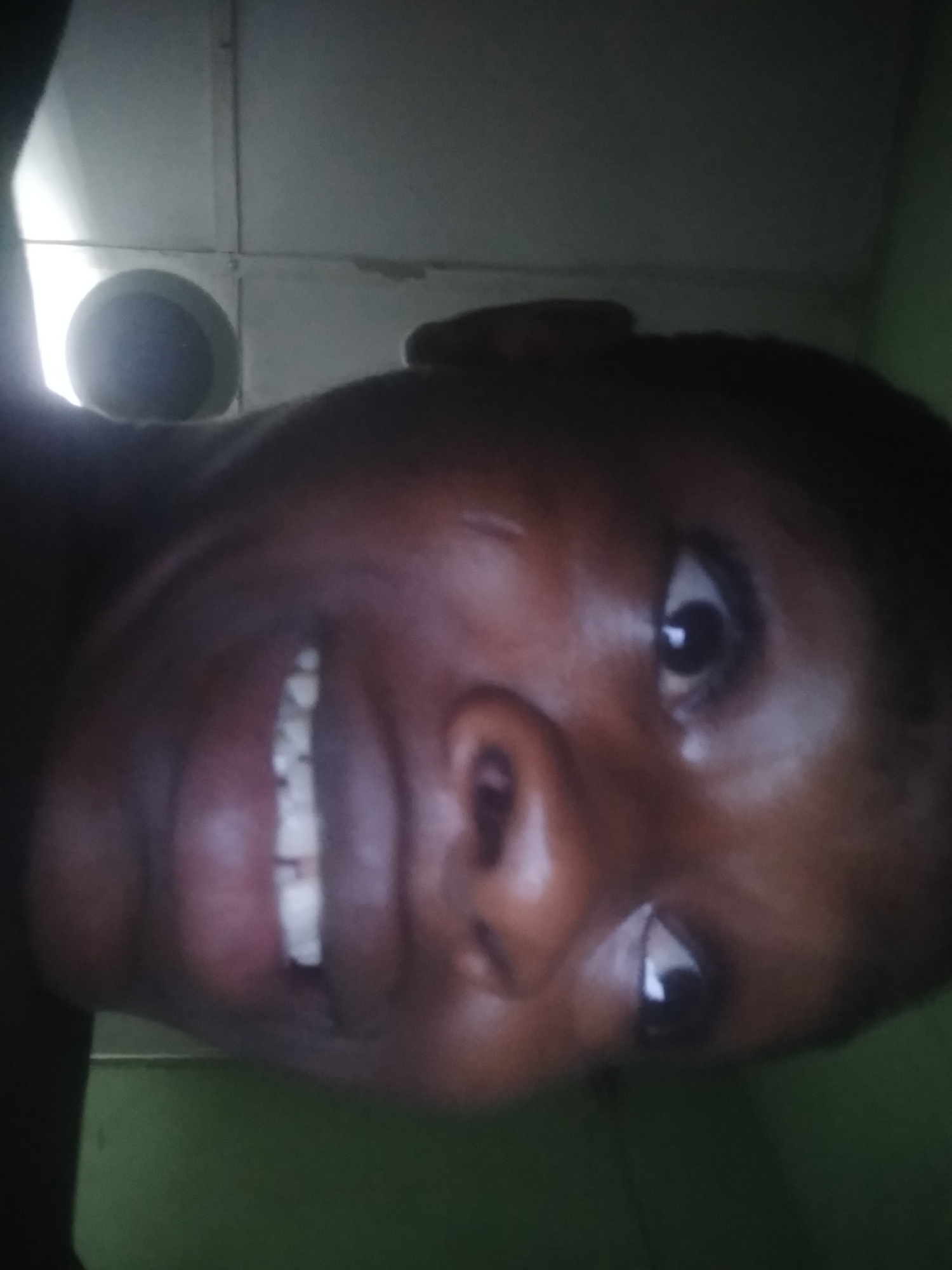 A close-up, tilted, low-angle view shows a person's face. The person has dark skin, and their teeth are visible in a smile. The background is indistinct, but parts of a ceiling and wall can be seen. The location is consistent with an indoor setting in Shangisha, Nigeria. Specific details beyond the subject's face and immediate surroundings are not discernible.