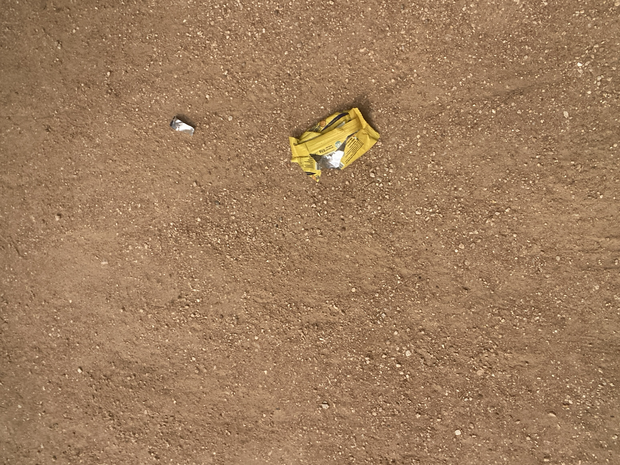 A close-up, overhead view of a dirt surface with scattered small pebbles and some litter. In the lower right quadrant of the image, a crumpled yellow wrapper from a snack or candy bar lies on the ground. To the upper right of the wrapper, a small piece of silver foil is visible. The environment appears to be an unpaved outdoor area.