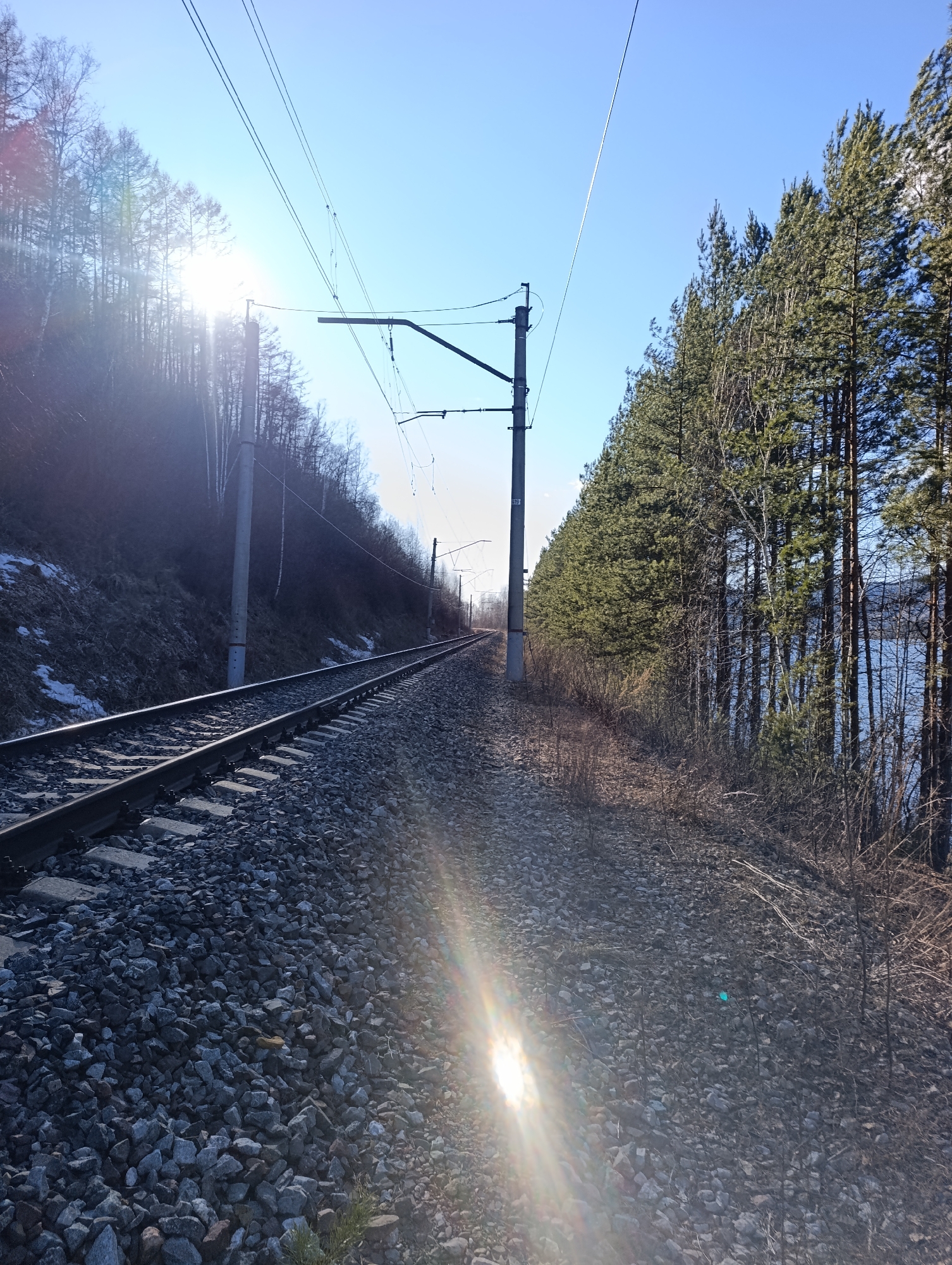 The image depicts a section of the Trans-Siberian Railway near Krasnoyarsk, Russia. The railway track extends into the distance between a wooded hillside on the left and a stand of trees on the right, beyond which a body of water is visible. Overhead power lines run parallel to the track, supported by metal poles. The ground is covered with gravel, and the sky is clear.