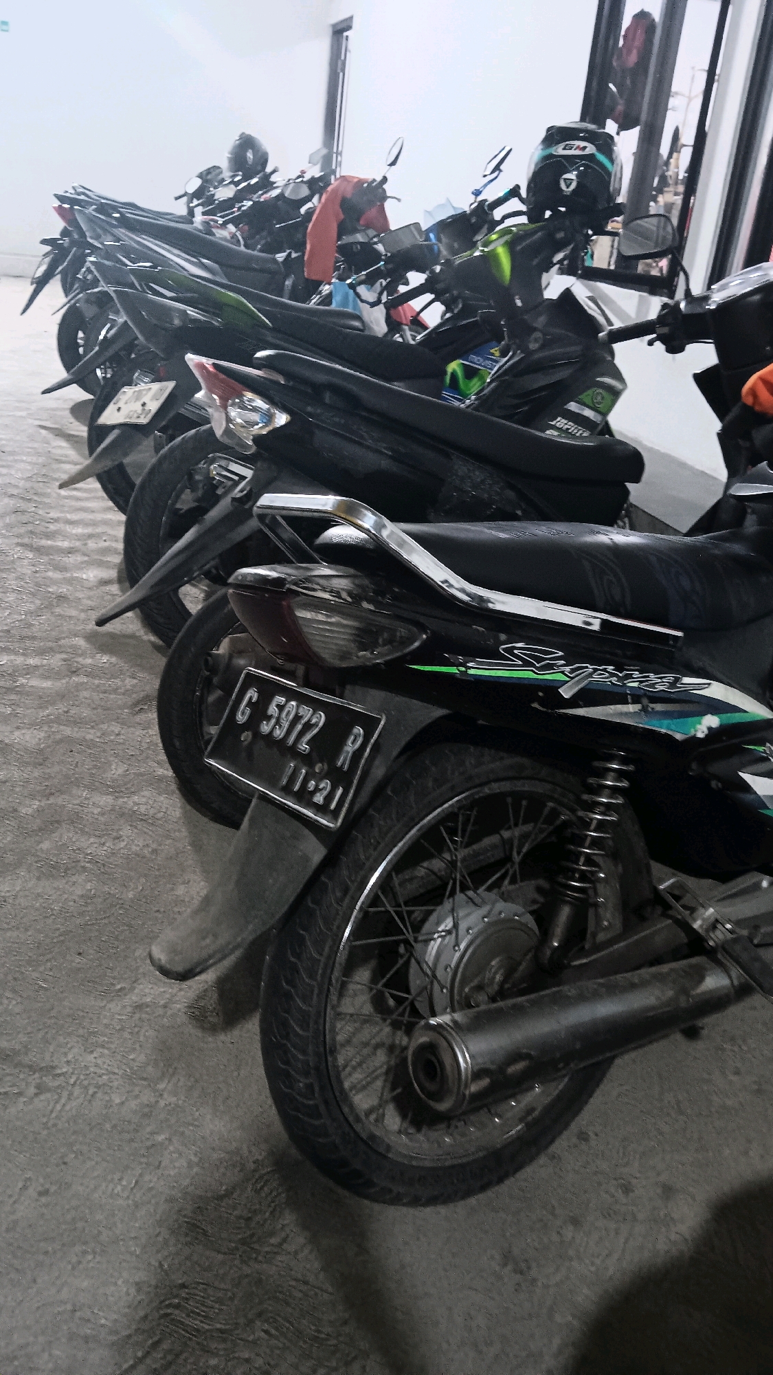 A line of parked motorcycles is shown in what appears to be a covered outdoor area. The motorcycles are primarily black with some metallic accents. In the foreground, a motorcycle with a license plate reading "G 5972 R" and "11-21" is prominently displayed, showing its rear wheel and exhaust pipe. A blue and black helmet rests on the handlebars of a motorcycle further back in the frame. The ground is a textured concrete surface, and the background shows a wall and some vertical elements, suggesting a building. The lighting is somewhat dim, casting shadows on the ground. The scene is likely a parking area for motorcycles in a public or residential space within the city of Pangebatan, Indonesia.