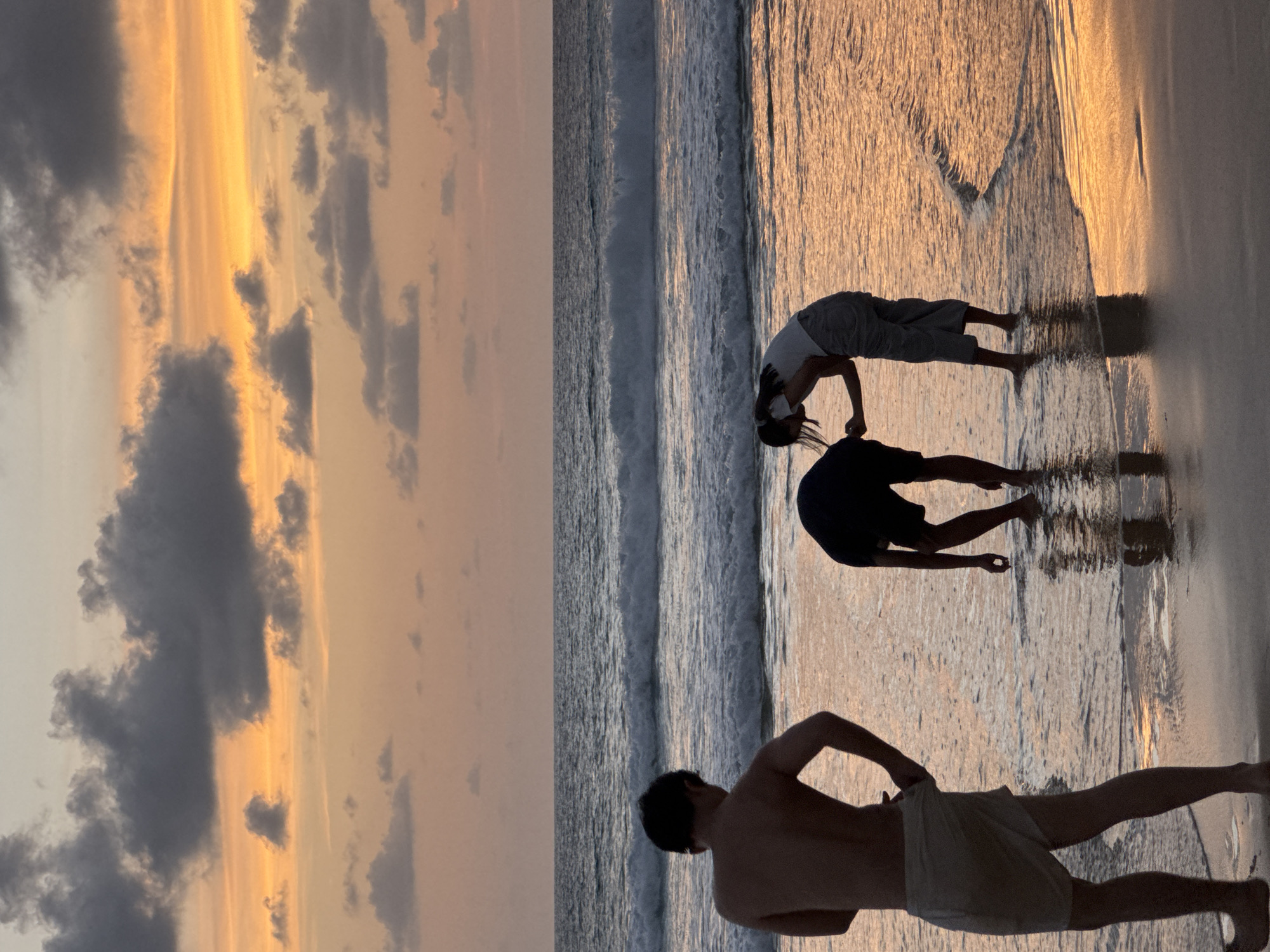 In Karon, Thailand, at sunset, three individuals are silhouetted on a beach. The sky is a gradient of orange and pink, with dark clouds scattered across it. The ocean has moderate waves, and the water laps at the shore. The person on the left, facing away from the camera, is wearing shorts. Two other figures, one wearing dark shorts and the other light pants and a white top, are interacting in the shallow water. The sand reflects the colors of the sunset.