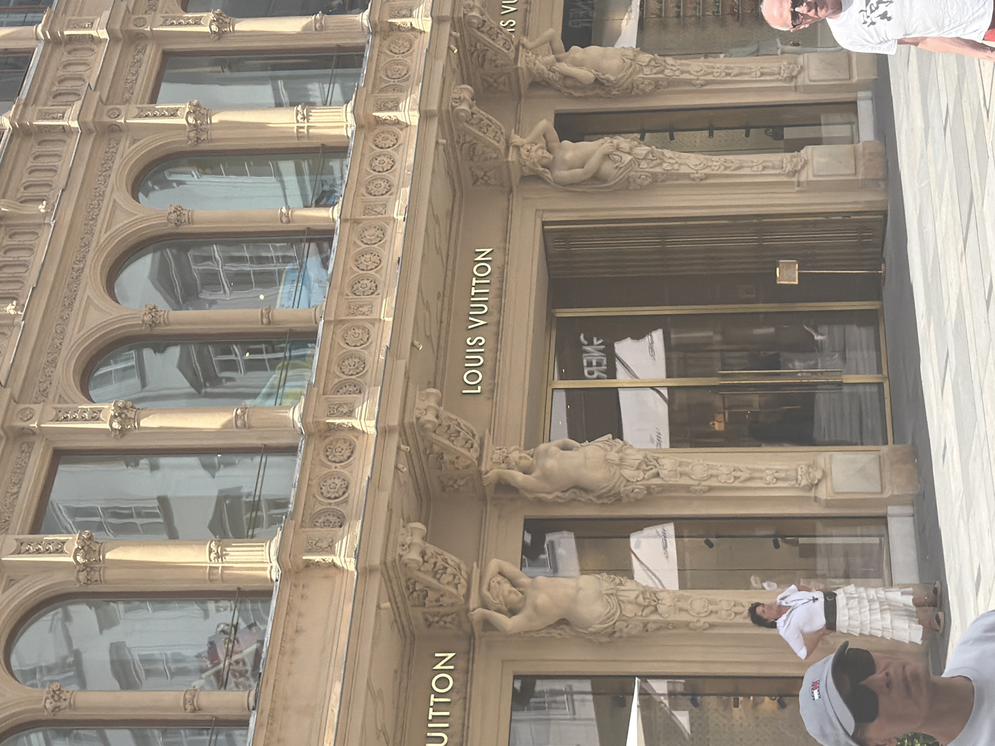 This media file captures a close-up, slightly angled view of the Louis Vuitton store facade in Vienna, Austria. The building's architectural style is ornate, featuring classical details like arches, decorative carvings, and sculptures of reclining figures adorning the upper levels. The facade is primarily light beige stone.
The activity taking place is not explicitly clear, but the presence of people suggests it's an active storefront, likely during business hours. One person, a man wearing a white t-shirt and sunglasses, is partially visible in the upper right corner, looking towards the camera. Another person, wearing a white ruffled skirt and white top, is standing near the entrance on the right side of the frame, looking away. A third person's head and shoulders are visible in the bottom right, wearing a grey baseball cap and sunglasses, also looking away.
Notable details include the large arched windows on the lower floors, which reflect the surroundings, and the prominent "LOUIS VUITTON" text in gold lettering on the facade. The lighting suggests it's daytime, possibly a sunny day given the reflections. The overall impression is one of a luxury retail environment combined with historical architecture.