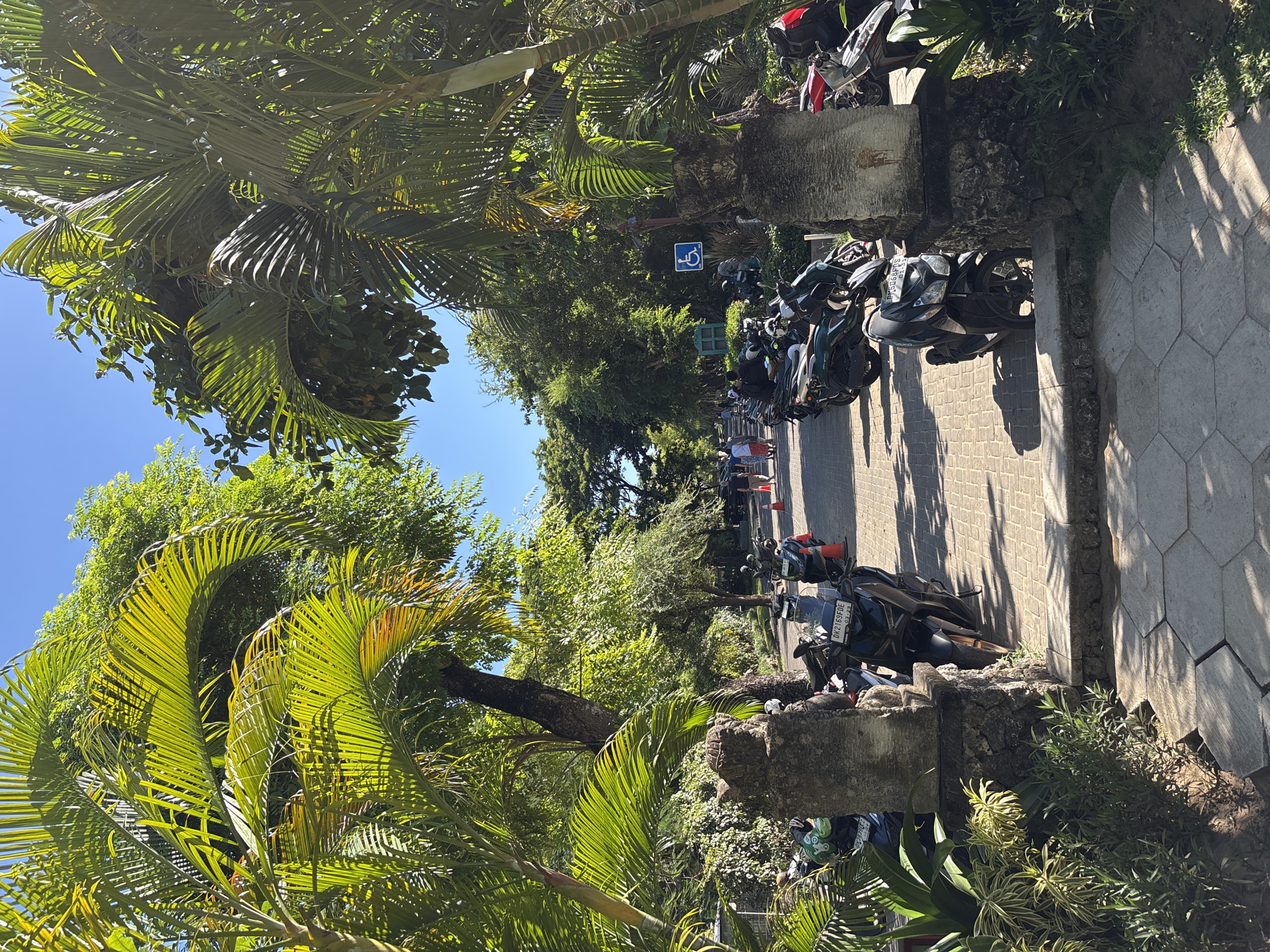 The image shows a street scene in Nusa Dua, Indonesia, with numerous motorcycles parked along the edge of the paved road. The area is shaded by lush, tropical vegetation, including palm trees. A blue handicapped parking sign is visible. People are walking in the distance. The road is constructed of rectangular paving stones. Concrete barriers separate the parking area from the vegetation.