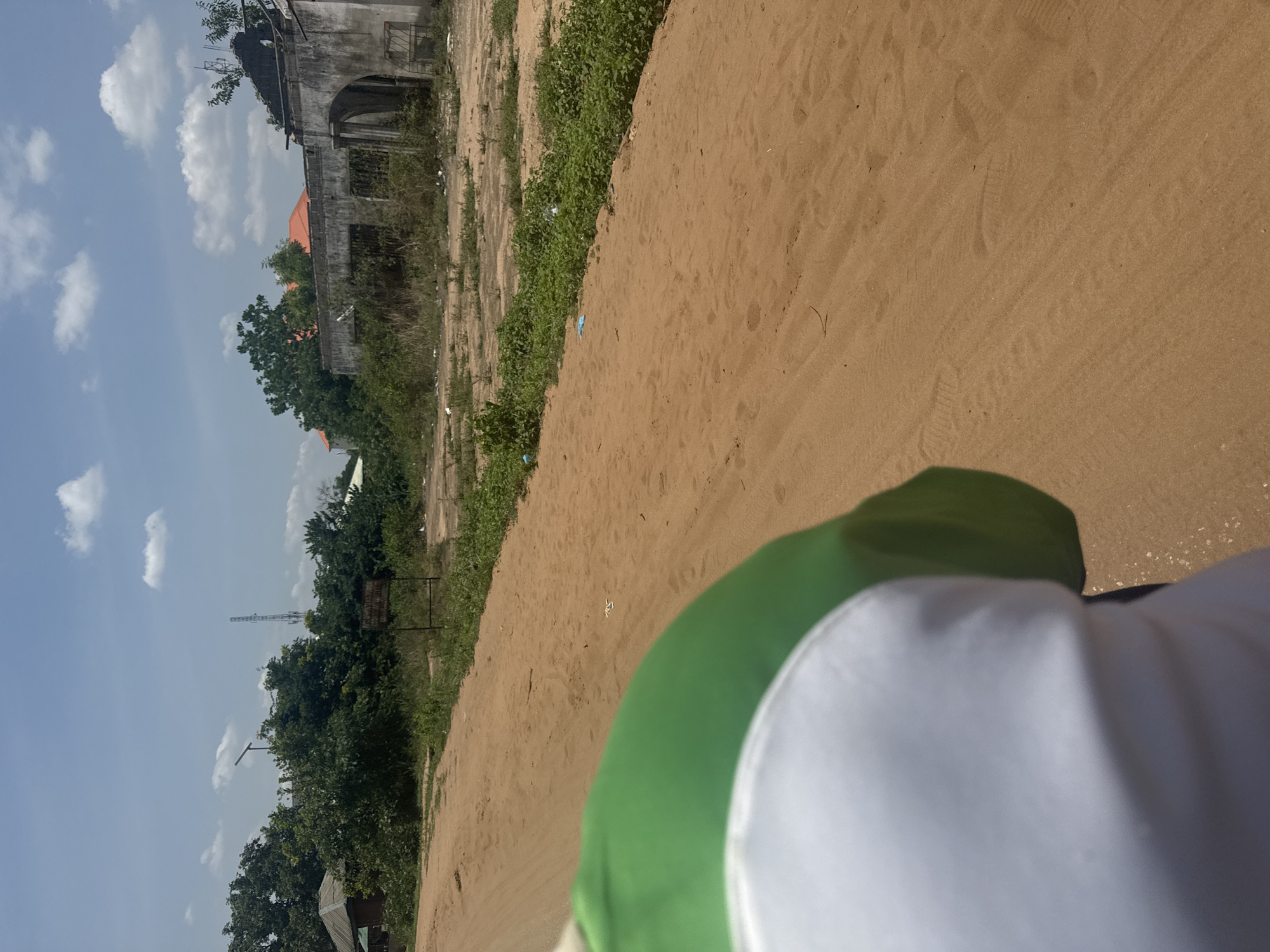 The image presents a view of a sandy area or path in Ihiagwa, Nigeria, with vegetation and trees lining one side. An aged, weathered building is visible in the background. A partially visible object in the foreground appears to be green and white. The sky is blue with scattered clouds. There is a tall communications tower visible in the background to the left.