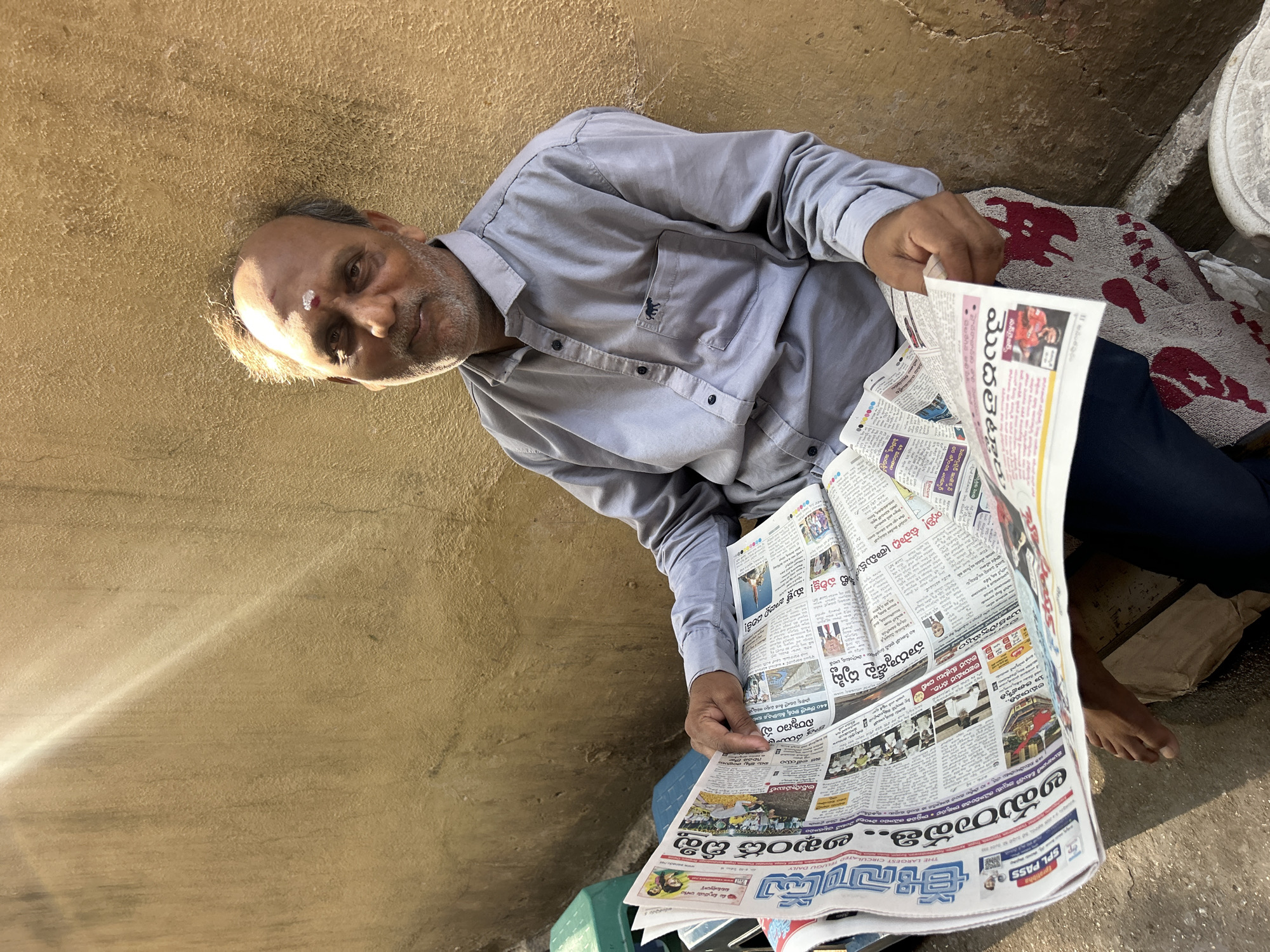 An elderly South Asian man in Puttaparthi, India, sits outside reading a Telugu newspaper. He wears a light blue long-sleeved shirt, dark blue pants, and a white tilak on his forehead. The newspaper is spread out in front of him, resting on his lap and a green container. He sits on the ground next to a blanket with red patterns. The background consists of a rough, tan-colored wall. The light indicates it is likely daytime.