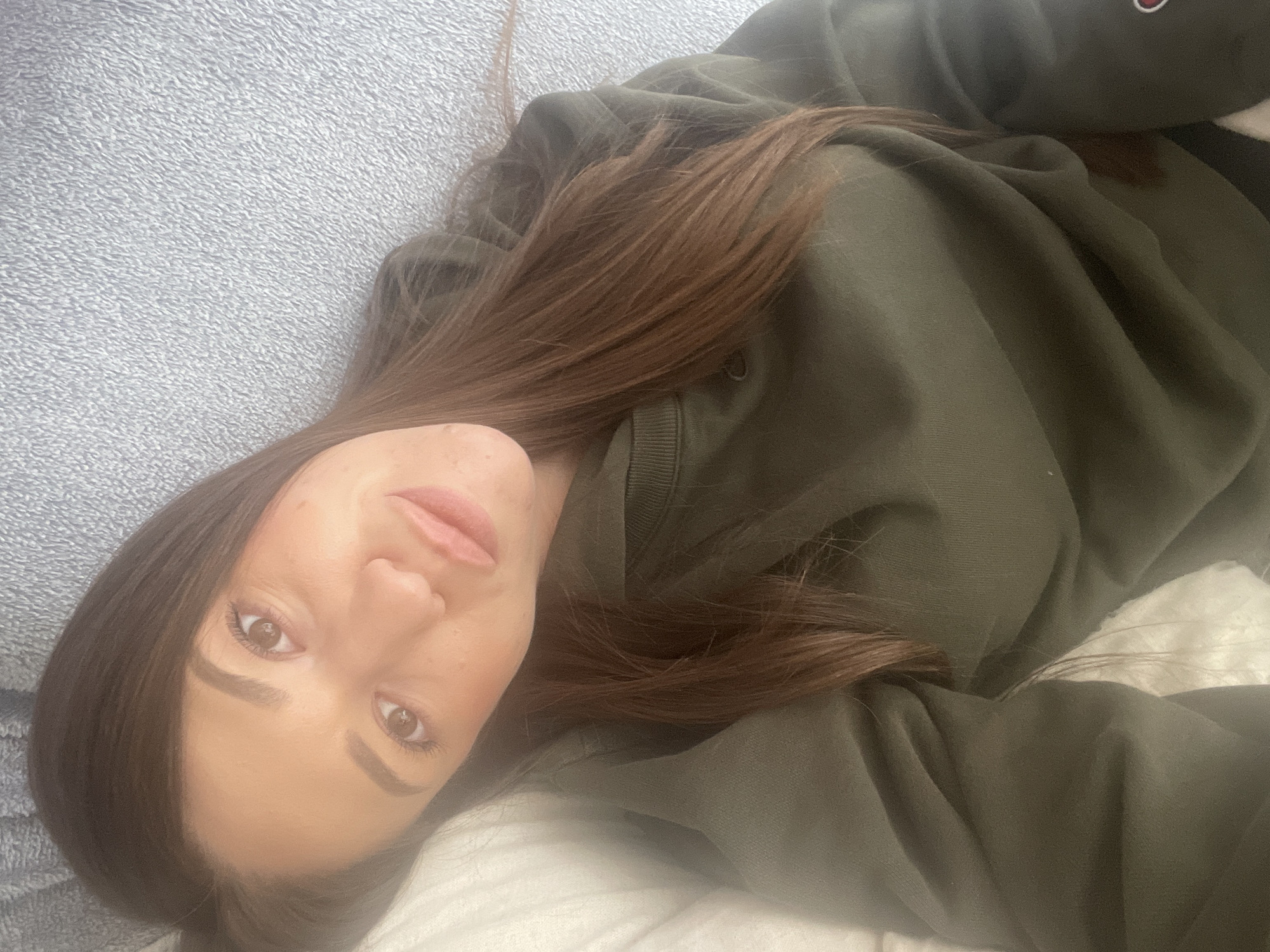 A woman lies on a light blue, textured surface. She has long, straight, brown hair that falls around her face and onto an olive green sweatshirt she is wearing. Her face is visible, showing brown eyes, fair skin, and neutral-toned lips. A white object is partially visible to her right. The setting appears to be indoors, possibly a bedroom or living area. The location in the city of Sofia, Bulgaria cannot be determined from the visual information provided.