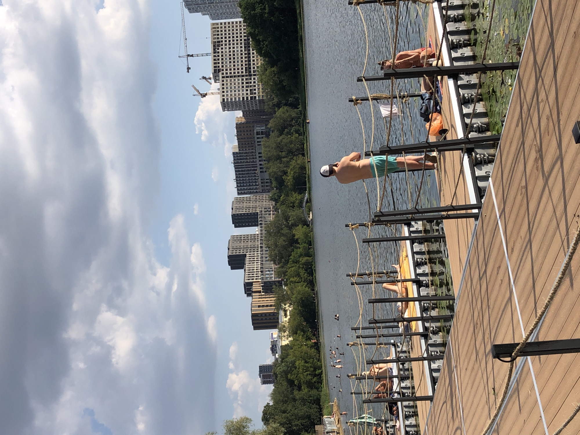 The image depicts a sunny day at a waterfront recreational area in Khimki, Russia. In the foreground, a wooden pier extends into a calm body of water. Several people are visible on floating platforms extending from the pier, sunbathing and relaxing. One man is standing on the pier, facing away from the camera, wearing a cap and turquoise shorts. In the middle ground, the water is populated with numerous swimmers. The background features a dense line of green trees, behind which a cityscape of modern apartment buildings and construction cranes is visible. The sky is partly cloudy.