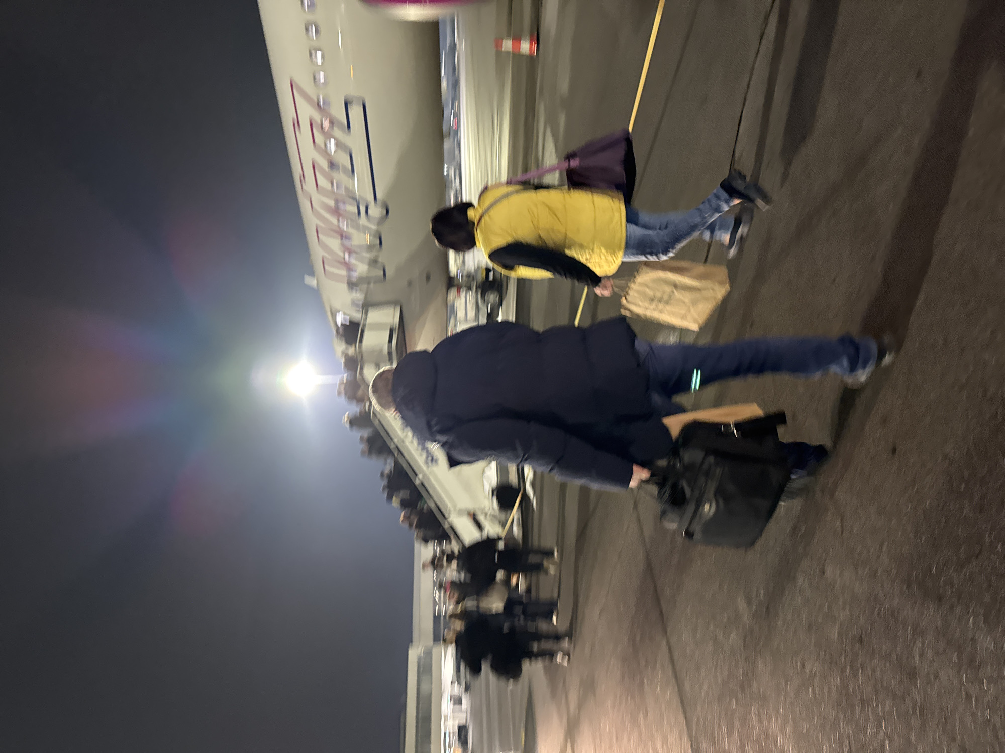 The image depicts passengers deplaning a Wizz Air aircraft at night. The location is likely Sofia Airport in Bulgaria, given Wizz Air's presence there. Passengers are disembarking via a staircase and walking on the tarmac. Individuals carry luggage and personal items. The environment is dark, illuminated by the plane's lights and airport lighting.