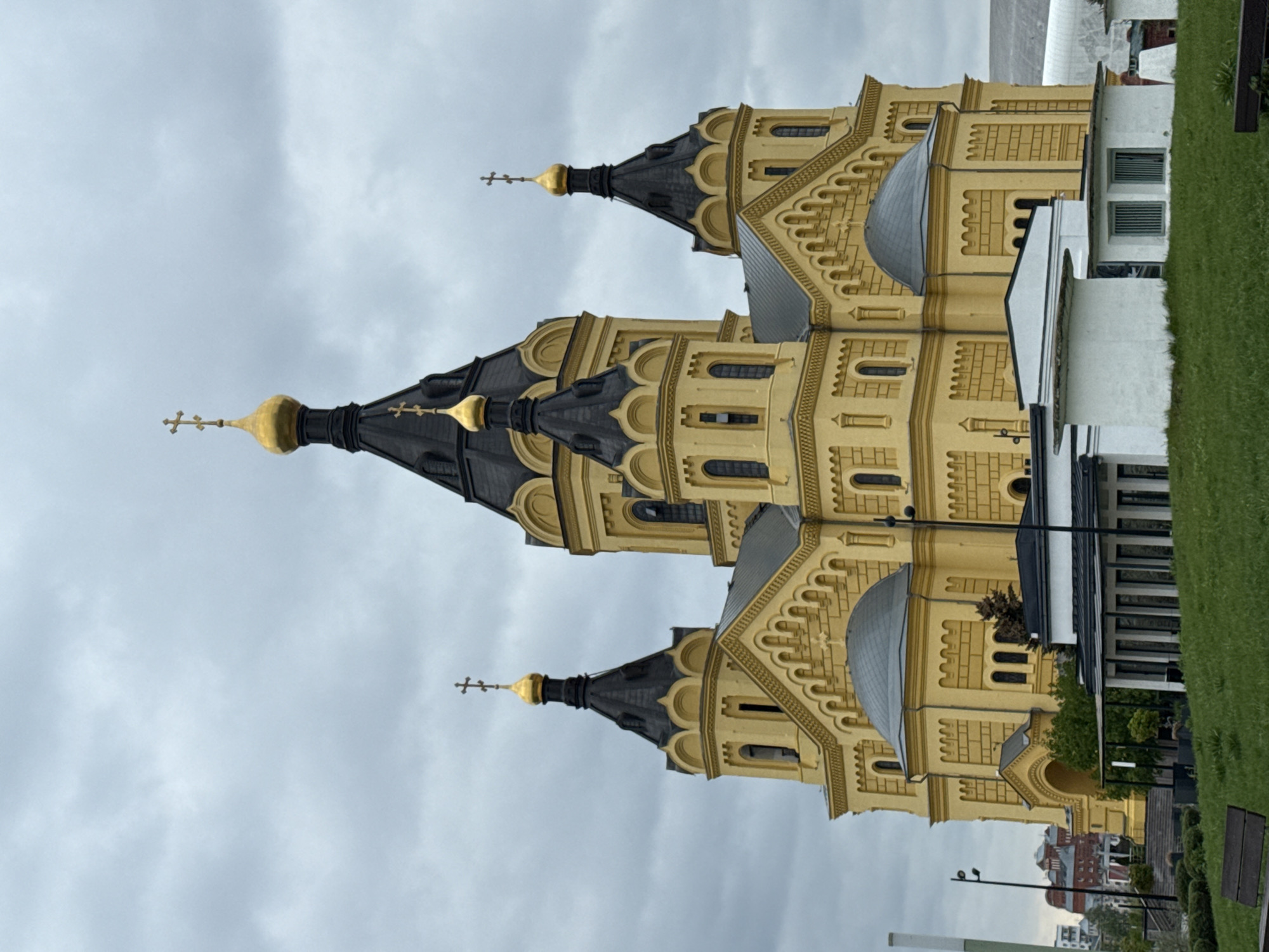 The image depicts the Alexander Nevsky Cathedral in Nizhny Novgorod, Russia. The cathedral is predominantly yellow with dark gray domes topped with golden crosses. The architecture is intricate with numerous arched windows and decorative moldings. A portion of the surrounding cityscape is visible, including a white building adjacent to the cathedral and distant residential structures. The sky is overcast with gray clouds, suggesting a cloudy day. Grass and some vegetation are also visible, indicating the cathedral is situated in a park-like setting.