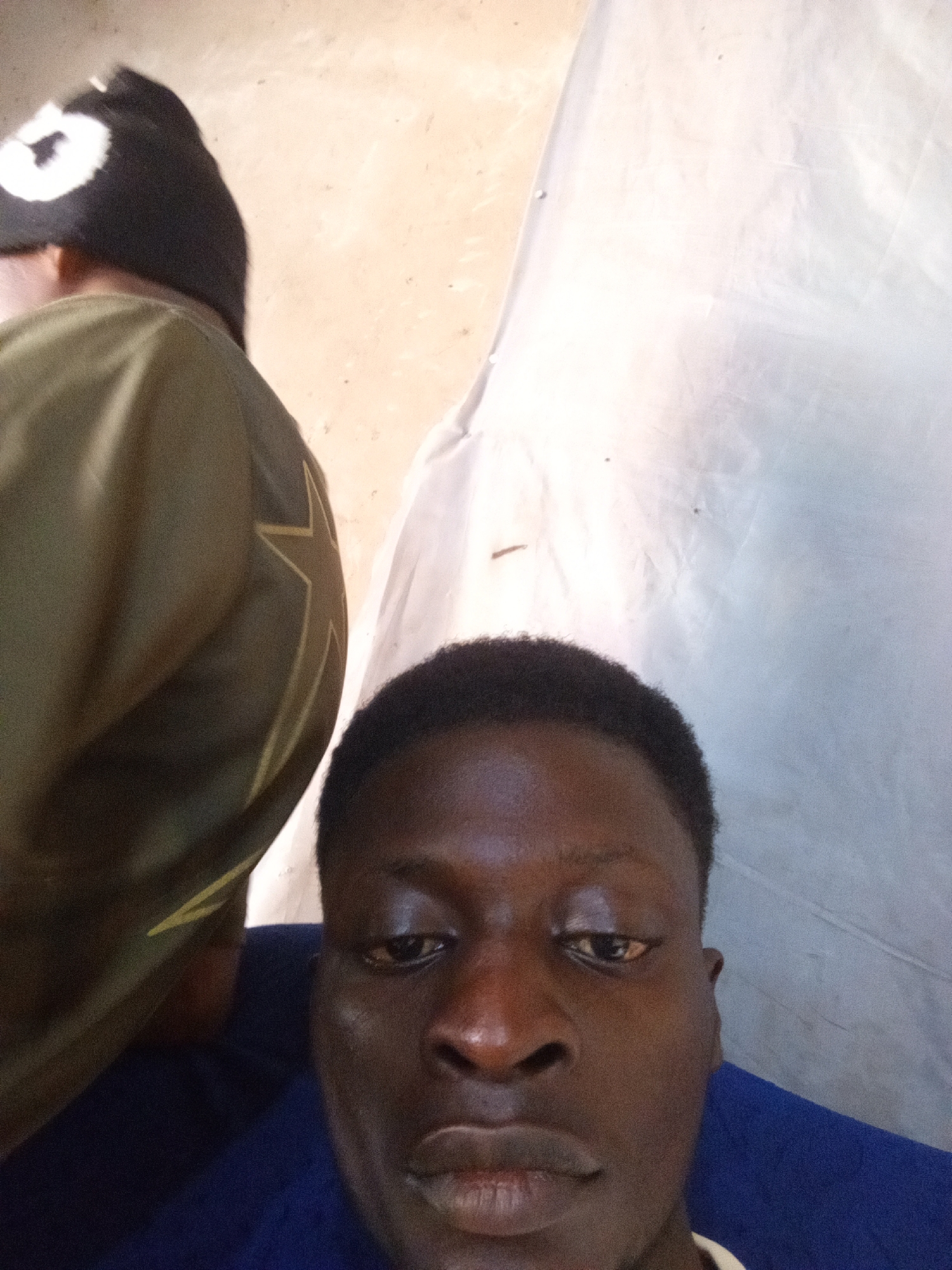 The image shows a close-up of a young Black man's face, looking directly at the camera. He has dark skin, short dark hair, and is wearing a dark blue shirt. To his left, another person's shoulder and back are visible, wearing a green garment with a yellow star design. This person is also wearing a black hat with a white circular design. The background is a light beige wall and a white cloth. The setting appears to be indoors, possibly a room in Nyabe, Nigeria.