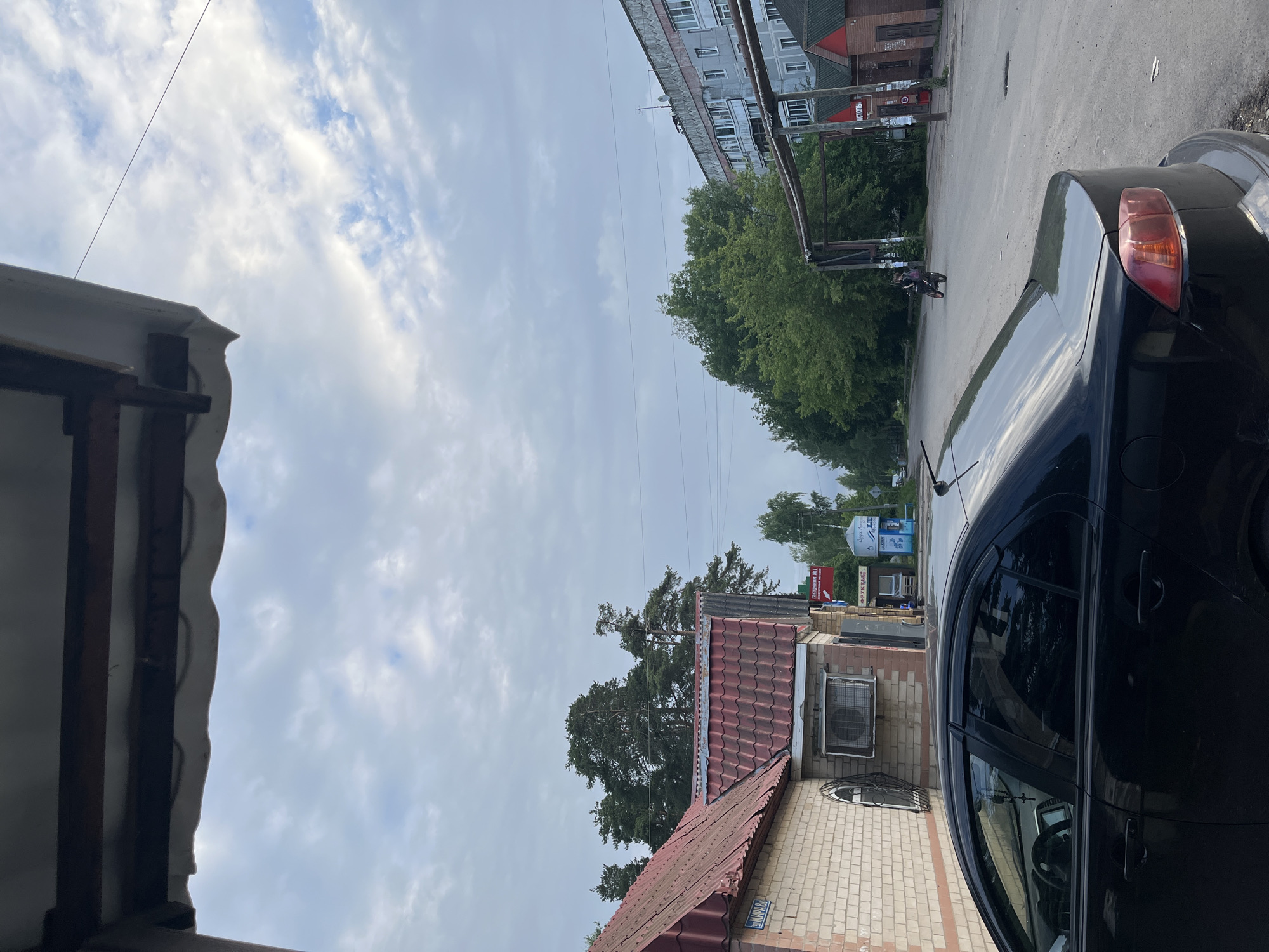 In Никольское, Russia, a black sedan is parked on a paved road. The car's interior is visible through the tinted windows, with a cross hanging from the rearview mirror. In the background, a person rides a motorcycle on the road. Several buildings line the street, including a brick building with a red tiled roof and a multi-story apartment building. Trees and a cloudy sky complete the urban landscape.