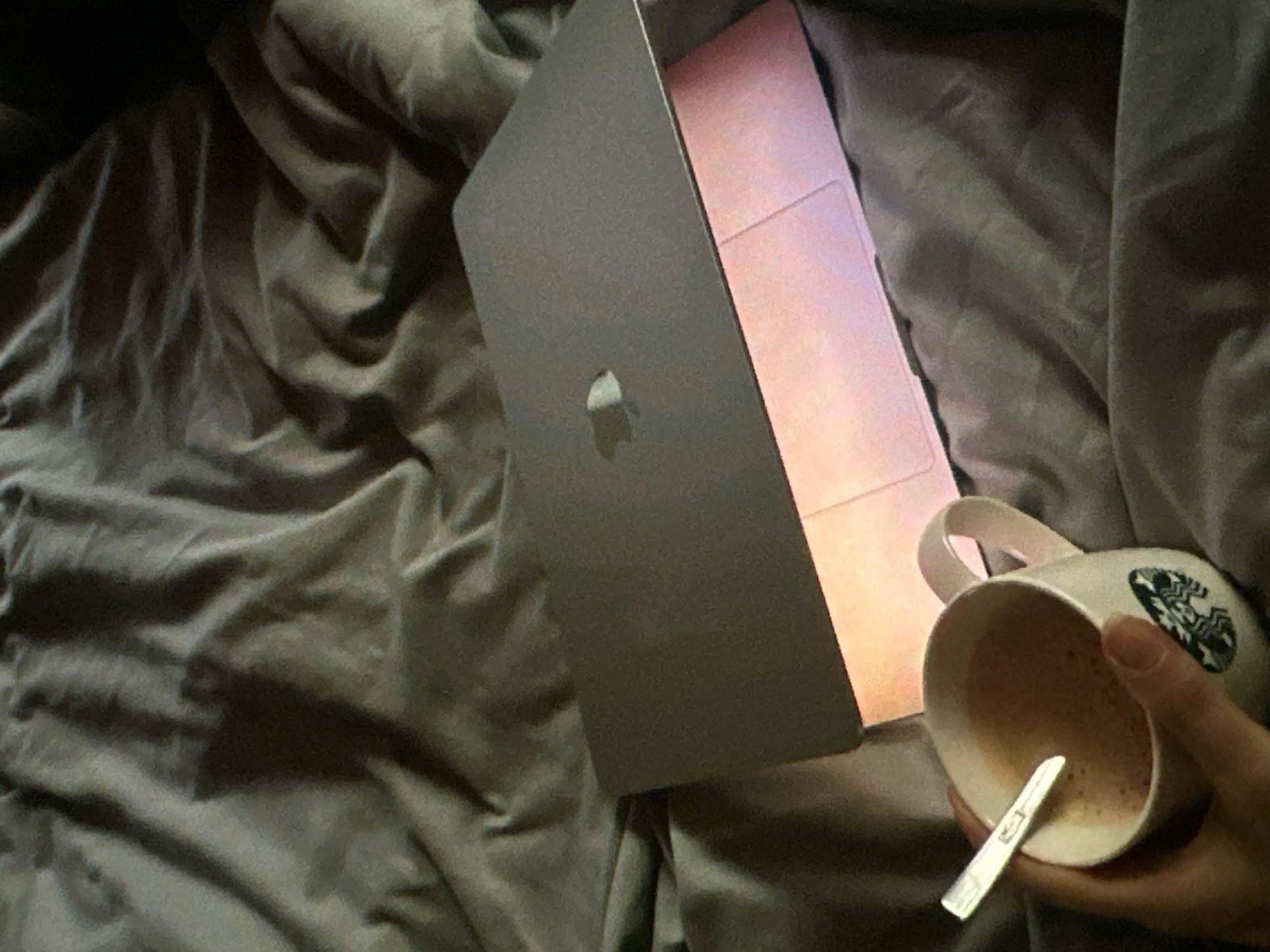 A grey Macbook is partially open on a grey crumpled bedspread. A Starbucks mug containing coffee with a metal spoon rests beside the laptop, held by a human hand. The location is not discernible from the image.