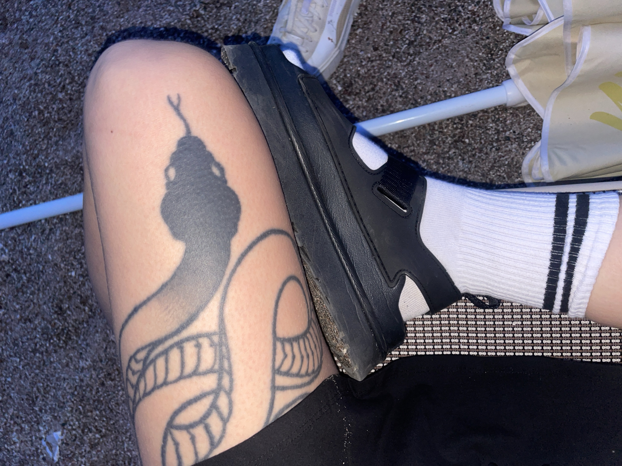 The image presents an overhead view focusing on a person's leg with a prominent snake tattoo. The tattoo depicts a stylized serpent with a distinct head and scaled body. A black sandal is positioned partially on the leg, obscuring part of the tattoo. The person wears a white sock with three black stripes at the top. Other details include a glimpse of a white shoe, an umbrella support pole, and a woven mat surface.
The image was not taken in Rafailovići, Montenegro.