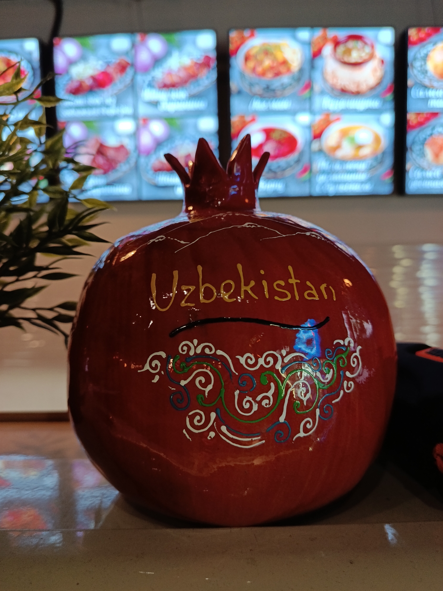 A red, decorative pomegranate with the word "Uzbekistan" written on it in gold paint sits on a light-colored surface. The pomegranate also features stylized, white, green, and blue decorative swirls. Behind the pomegranate is a plant with small, dark green leaves on the left and a series of backlit panels displaying food images. The location is Moscow, Russia.