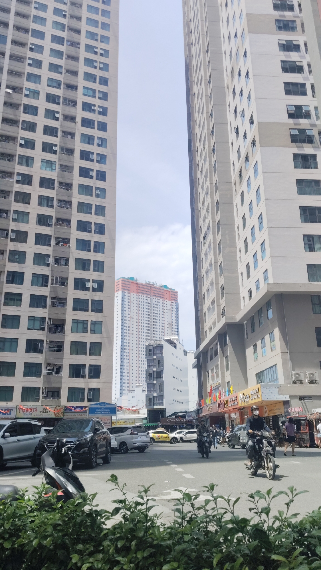 The scene is a street in Bac Nha Trang Ward, Vietnam, framed by tall buildings on either side. In the midground, there is an intersection with cars, motorbikes, and pedestrians. One person is riding a motorbike and wearing a mask. There are shops and restaurants visible along the street. In the background, another tall building is seen through the gap between the buildings flanking the street. Lush greenery is prominent in the foreground.