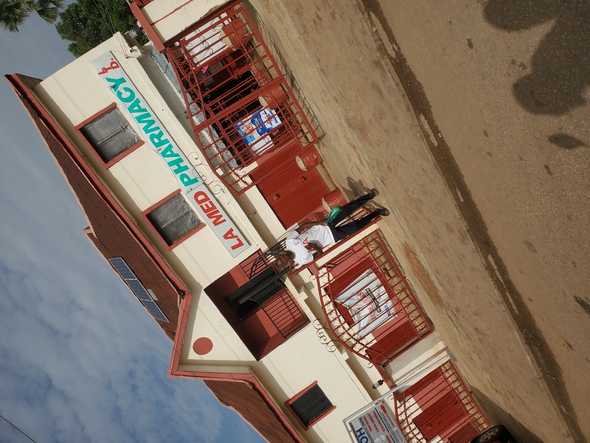 In Bukuru, Nigeria, a man stands in front of a beige building with a red-tiled roof. The building has a sign that reads "LA MED PHARMACY LTD." in large, green letters. To the left of the pharmacy, there is another sign for a "DIAGNOSTIC CENTRE." Red metal gates are in front of the entrance, with posters visible through the bars. A man, wearing a white t-shirt and dark pants, is casually posed, holding a green bag. The street in front of the building is paved with asphalt and shows signs of wear. The sky is partly cloudy.