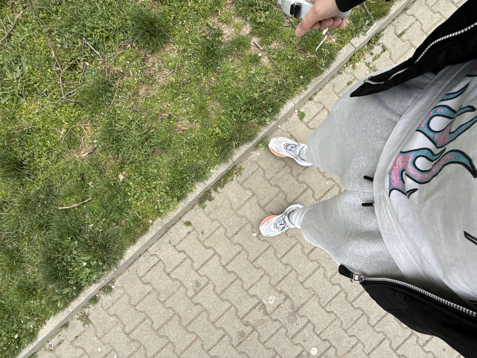 The scene depicts a high-angle view looking down at a person standing outdoors in Stara Zagora, Bulgaria. The subject's lower torso, legs, and feet are visible. The individual is wearing a white t-shirt featuring a pink and blue graphic design, light grey sweatpants, and white athletic shoes with orange and light blue accents. A black jacket is partially visible on the left, unzipped. The subject's right hand, visible in the upper right quadrant, holds a light grey electronic device. The ground comprises a grey interlocking concrete paver sidewalk on the left, which abuts a concrete curb. To the right of the curb is an unkempt strip of ground covered with green grass, sparse dirt patches, scattered dry twigs, and small white flowers.