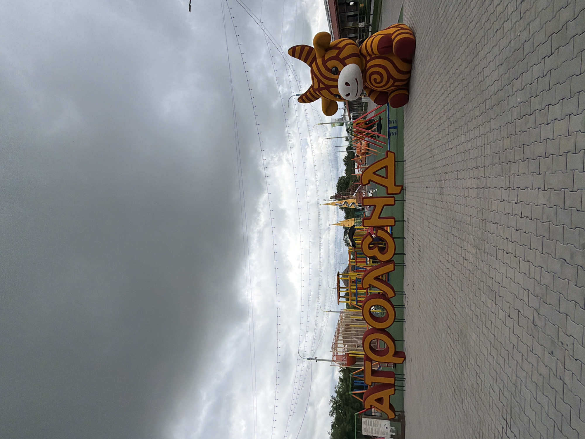 In Velykyi Khodachkiv, Ukraine, an outdoor recreational area features a large, orange and yellow stuffed bull with swirling patterns. To the left of the bull, large, red and yellow letters spell out "АГРОЛЕНД", which translates to "AGROLAND". Behind the letters is a colorful playground with swings and climbing structures. Overhead, strings of lights are suspended, and the sky is overcast with dark, grey clouds. The ground is paved with interlocking grey bricks. The overall scene suggests a family-friendly attraction or park.