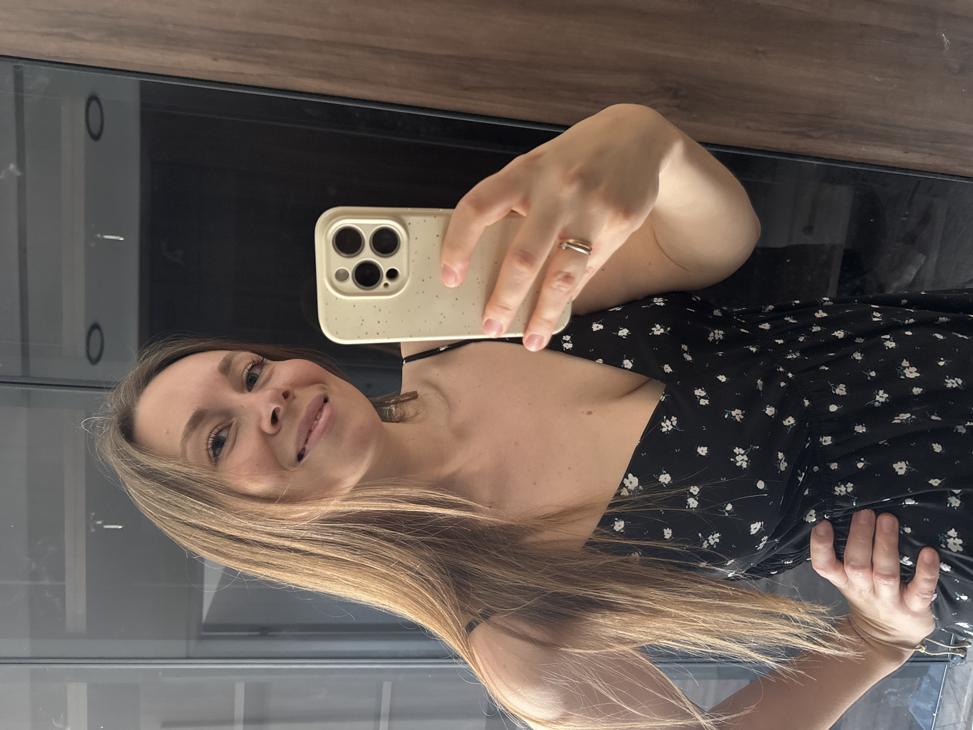 A woman is taking a selfie using her smartphone. She is wearing a black dress with small white floral patterns. She has long blonde hair and is wearing a gold ring on her ring finger. The background appears to be a modern interior with dark cabinetry. A reflection in the cabinetry reveals a possible window and door frame. The image does not contain enough information to identify the location as Budva, Montenegro.
