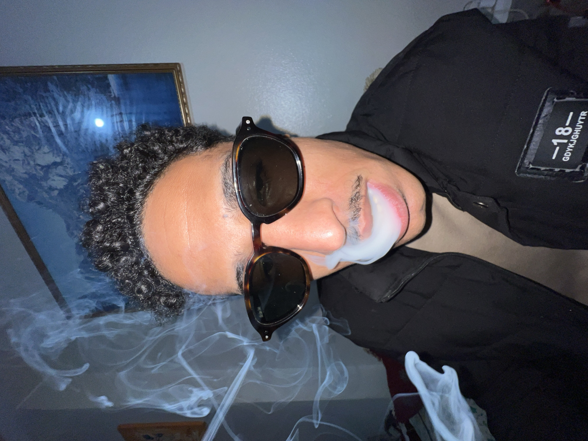 A person with dark curly hair and a mustache is wearing sunglasses and a black jacket. They are exhaling smoke from their mouth. The jacket has a patch on the upper right with the number "-18-" and the text "GDYKJGHUYTR". A framed picture hangs on the wall behind the person. The location is not specified, and it is not possible to determine if it is Tanger طنجة, Morocco.
