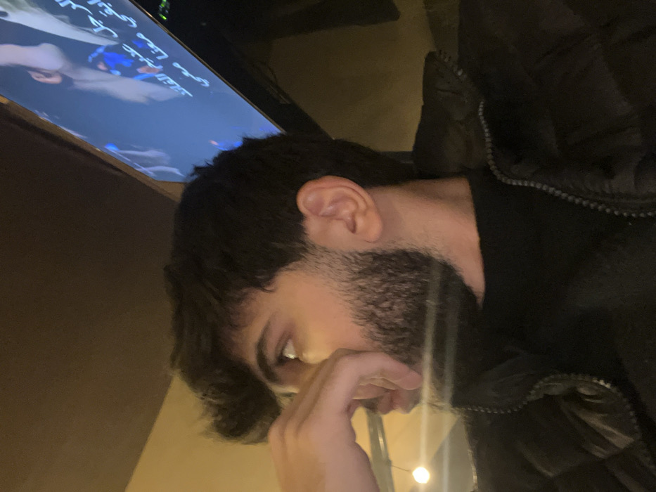 A dimly lit indoor scene captures a man gazing downward with a contemplative expression. His hand partially obscures his face, and a neatly trimmed beard is visible. He's wearing a black jacket. In the background, a television screen displays Arabic script. The lighting is soft, contributing to the subdued mood.