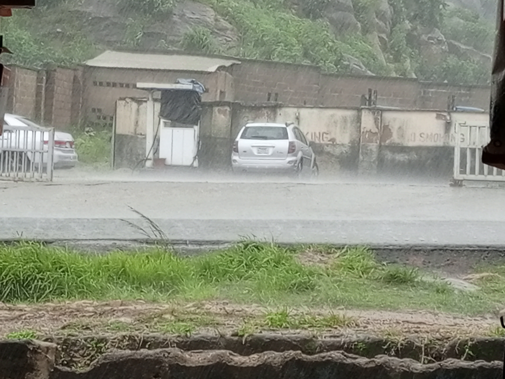 The image depicts a scene on Rukuba Road, Nigeria, during a heavy downpour. The road is wet and appears to be covered in a thin layer of water, indicating significant rainfall. In the background, a brick building with a corrugated metal roof is visible. Adjacent to it, a small structure with a white facade and a black tarp covering its upper section is present. A silver SUV is parked in front of these buildings, its rear facing the camera. To the left of the SUV, another silver car is partially visible behind a metal gate. A concrete wall with faded markings, including what appears to be "NO SMOKING," runs along the property. Lush green vegetation covers the hilly terrain behind the buildings. In the foreground, a strip of green grass is bordered by a concrete structure. The overall atmosphere is somber, dominated by the rain and the muted colors of the environment.