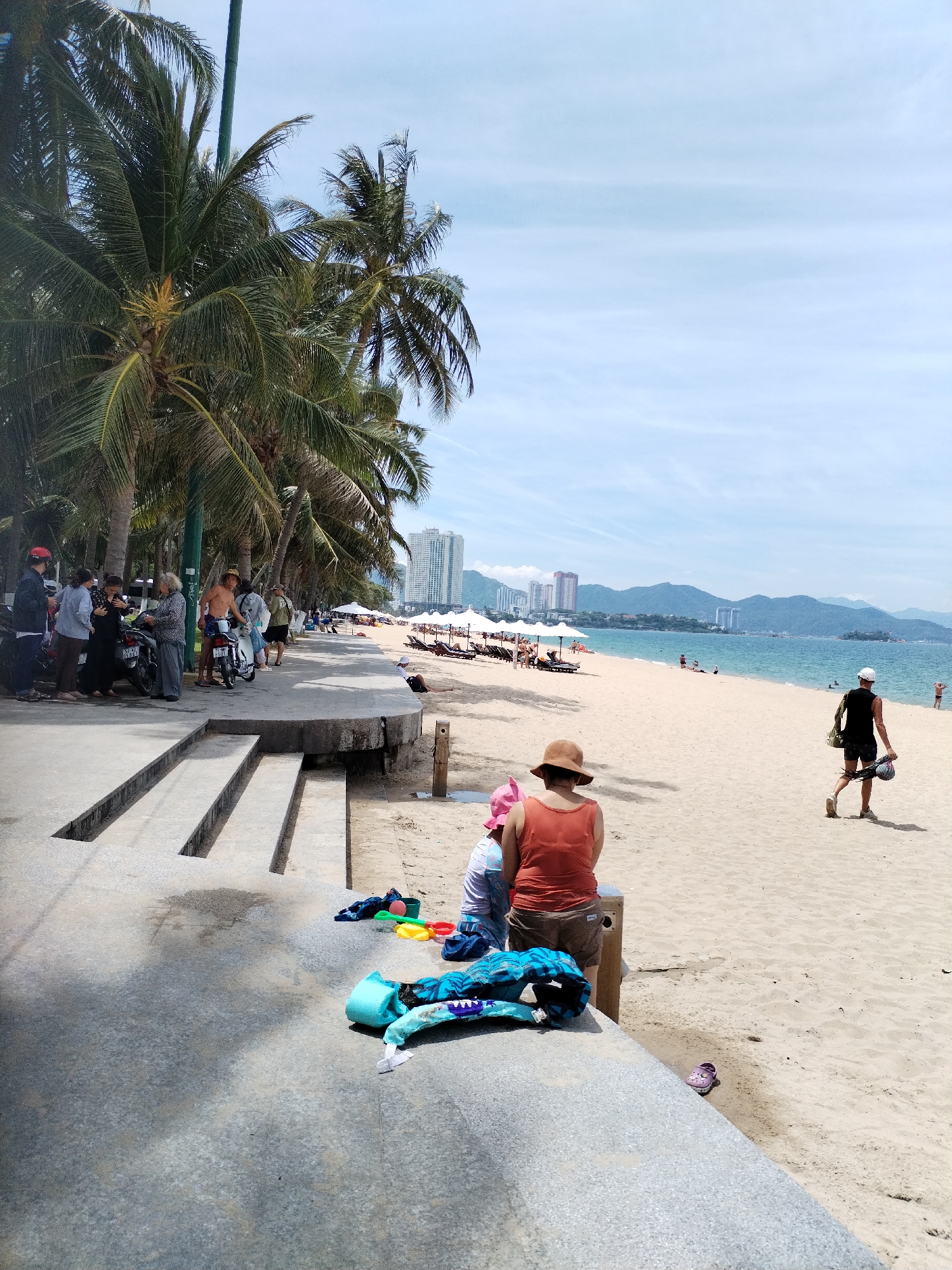 This image depicts a sunny day at Nha Trang Beach, Vietnam. The foreground features a concrete walkway with several sets of stairs leading down to the sand. Scattered on the walkway are children's beach toys, including a blue life vest, a green shovel, and colorful buckets.
Further into the scene, a sandy beach stretches out, lined with palm trees and white umbrellas. Several people are visible, including individuals walking along the shore, relaxing on beach chairs, and swimming in the ocean. In the middle ground, two women and a child sit on the edge of the concrete walkway, facing the beach. One woman wears an orange tank top and a wide-brimmed hat. Behind them, a man in a black tank top and shorts walks away from the camera, carrying a bag over his shoulder.
The background reveals the Nha Trang coastline with a backdrop of rolling hills and high-rise buildings. The water is a clear blue, with a few small boats visible on the horizon. The overall atmosphere is one of leisure and recreation.