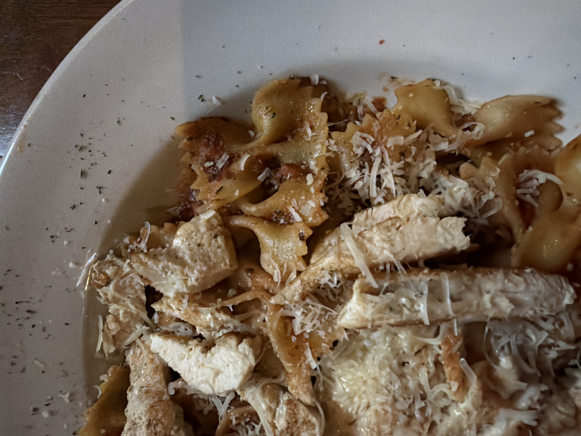 A top-down, close-up view shows a partially consumed plate of pasta with chicken and cheese. The pasta is farfalle, commonly known as bow-tie pasta, and appears to be coated in a sauce. Chunks of chicken are scattered throughout the pasta. Shredded cheese is sprinkled on top of the pasta and chicken. The plate is white and has a slightly visible rim on the left side of the frame. The image appears to be taken indoors. The image shows a dish likely served at a restaurant in College Station, United States of America. The photograph shows a pasta dish, specifically featuring farfalle pasta, chicken, and cheese. The ingredients are arranged on a white plate and captured with a close-up perspective. The focus is on the textures and details of the food.