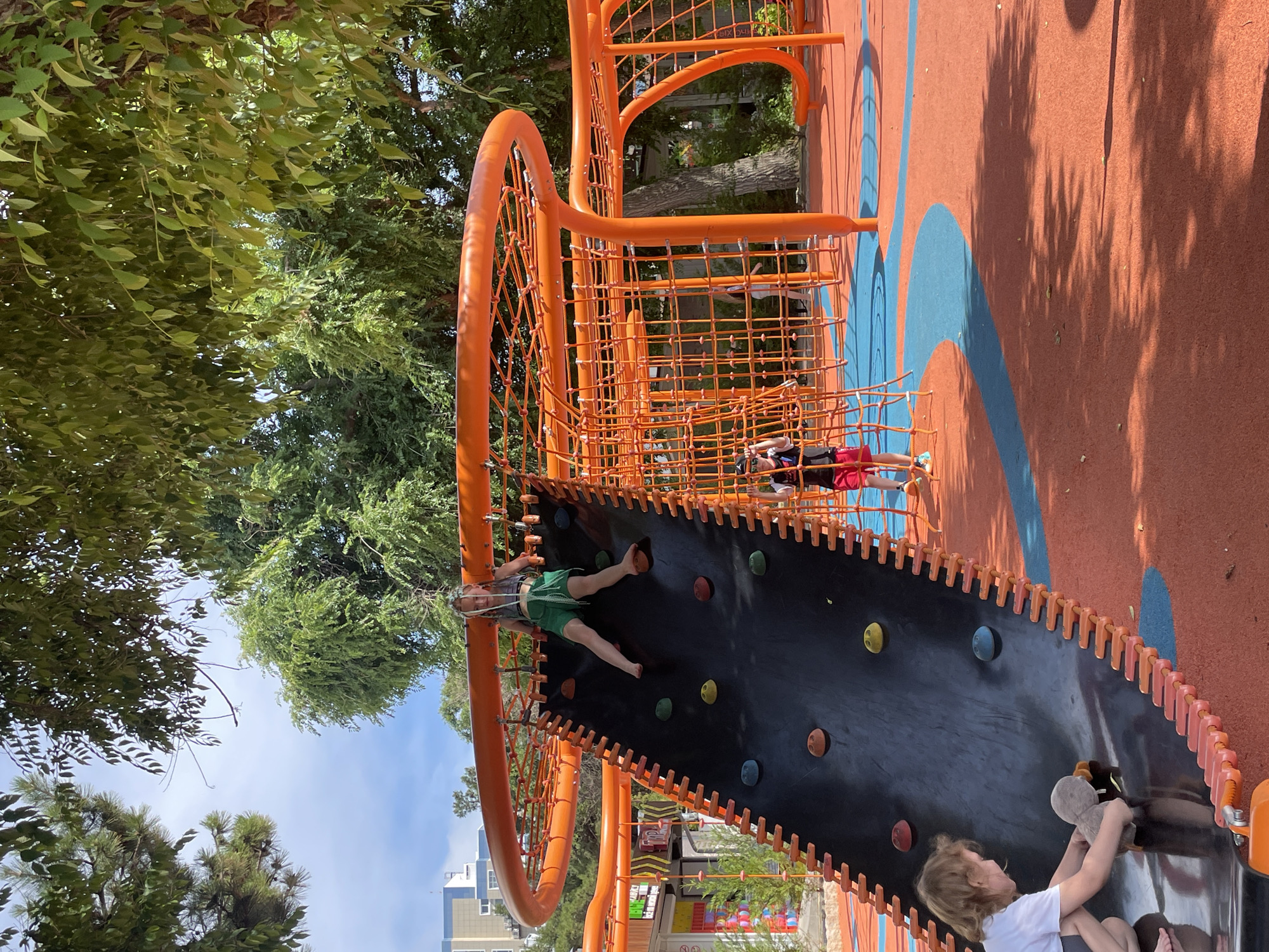 A group of children are playing on an orange and black playground structure in Anapa, Russia. The playground has a climbing wall with colorful grips and a net climbing area. One child is on the climbing wall, wearing a green shirt and shorts. Another child is in the net climbing area, wearing a red shirt and black shorts. A third child is at the bottom of the climbing wall, holding a stuffed animal. The ground of the playground is orange with blue curved lines. Trees with green leaves surround the playground. The sky is blue with some clouds. The lighting suggests it is daytime. The children appear to be enjoying themselves, with smiles visible on some of their faces. The overall impression is a cheerful and active outdoor scene. There is no visible text in the image.