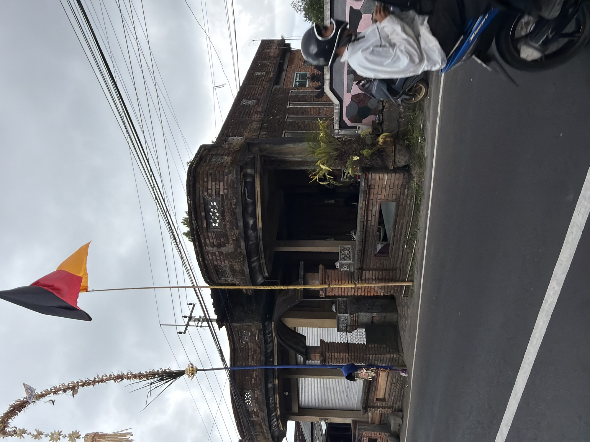 In Kintamani, Indonesia, a cloudy day is depicted. A man wearing a white shirt and a black helmet rides a blue motorcycle on a paved road. To his right, a building with weathered brickwork and an arched entrance stands prominently. A German flag flies from a tall pole in front of the building. Decorative elements, possibly part of a Balinese ceremony, hang from a tall pole on the left side of the scene. Power lines crisscross the sky above the building.