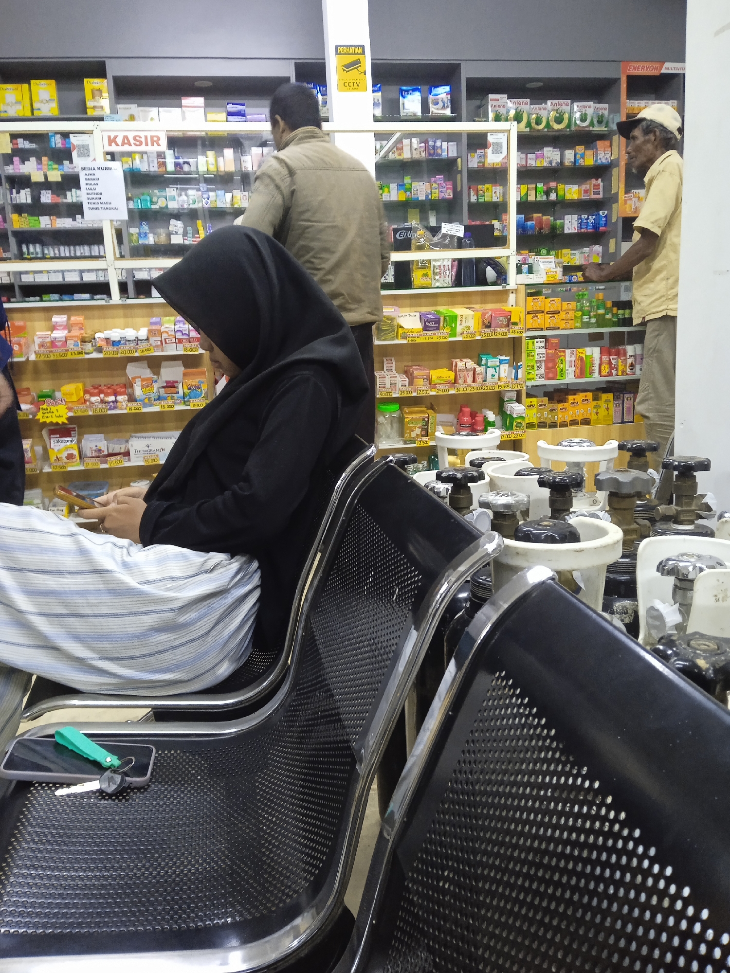 The image depicts an indoor scene within a pharmacy or drug store located in Kebonsari, Indonesia. In the foreground, a woman wearing a black hijab and striped light blue and white pants is seated on a black perforated metal chair, engrossed in her smartphone. Next to her, another identical empty chair holds a smartphone with a green keychain and a key. To the right, a cluster of white and black medical gas cylinders or oxygen tanks with large valve handles are visible.

The midground and background are dominated by tall shelves stocked with a wide array of pharmaceutical products, medications, and health supplements. Two men are present in this area. One man, seen from behind in a light brown jacket, stands near a section labeled "KASIR" (Cashier). To the right, an older man with a light yellow shirt and a beige baseball cap stands by another shelf, observing the products.

Notable details include various visible text. The "KASIR" sign is prominent. A sign above the cashier lists "SEDIA KURMA" (Available Dates) followed by types such as Ajwa, Bakari, Kolas, Lulu, Ruthor, Sukari, Tunis Madu, and Tunis Tangkai. Above the shelves, a yellow sign reads "PERHATIAN CCTV" (Attention CCTV). Numerous product brands like Anlene, Sakatonik, Theragran, Diabet (likely DiaBes), Darsi, Enervon Multivita, GPU, and Enfa are identifiable on the packaging. Many items display price tags, ranging from 5,000 to 140,000 units of currency, likely Indonesian Rupiah. The setting appears well-lit by artificial light, suggesting business hours.