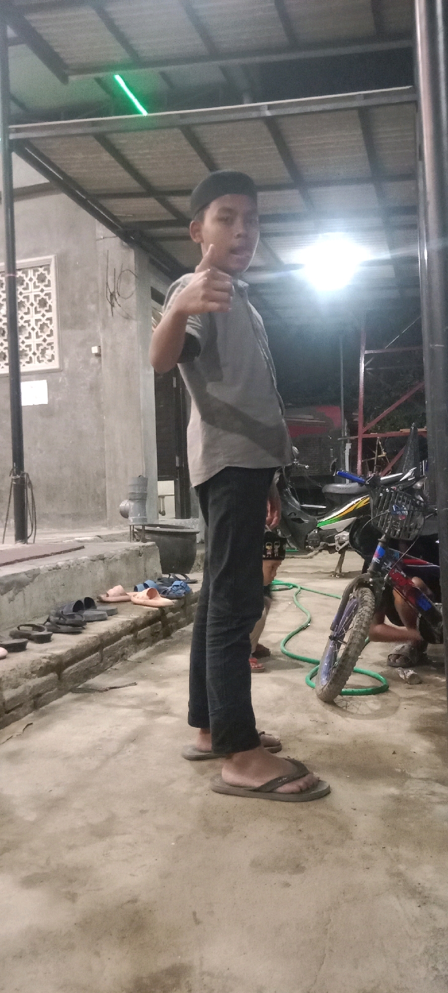 The image depicts a young person giving a thumbs-up gesture while standing in what appears to be an outdoor, covered area, possibly a porch or garage, located in Kras, Indonesia. The person is wearing a dark skullcap, a gray short-sleeved shirt, and dark trousers. They are also wearing flip-flops. In the background, a motorcycle is partially visible, and several pairs of sandals are arranged on a low concrete ledge to the left of the frame. The environment is somewhat dimly lit, with a bright light source in the background creating a flare. A green tube or hose is visible on the ground. The wall behind the young person has a decorative patterned window or vent.