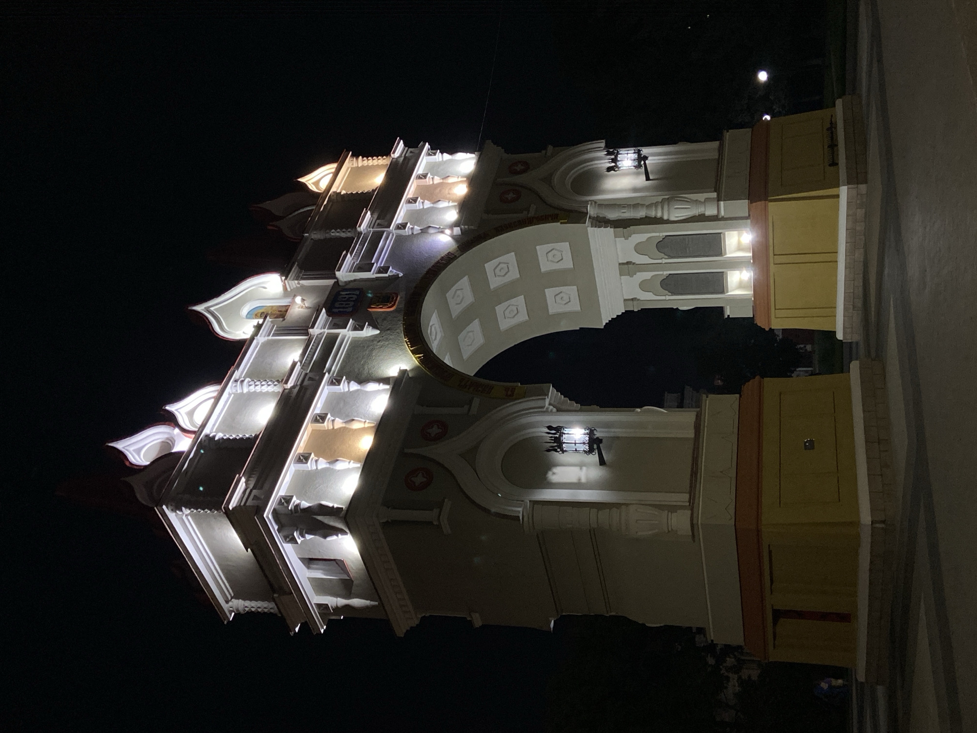The image shows a nighttime view of the Triumph Arch in Blagoveshchensk, Russia. The arch is illuminated by external lights, highlighting its architectural details. The structure is made of light-colored stone with decorative elements and a prominent archway. Above the arch, there are ornate structures topped with flame-like finials, some of which are also lit. A year, "1991", is visible on the facade. Two traditional lanterns are mounted on the sides of the arch's pillars. The ground in front of the arch appears to be paved. In the background, the night sky is dark, with a few distant lights from the city and trees silhouetted against the darkness. A person is faintly visible on the left side of the image, near the base of the arch, and another faint light source is visible in the far right background.