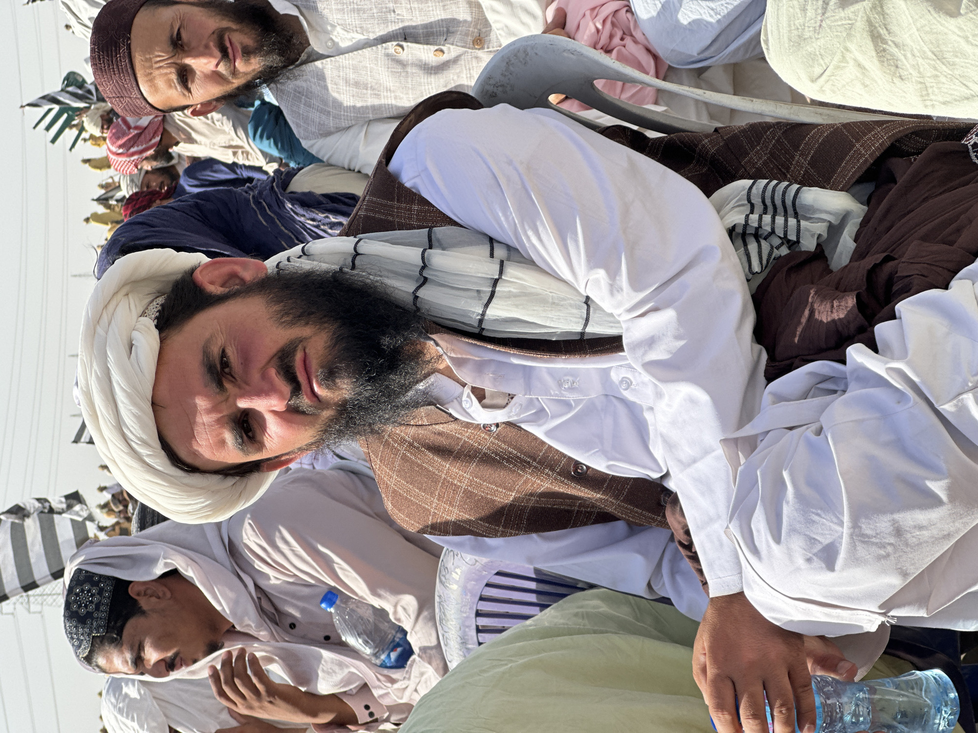 An outdoor scene in Pishin, Pakistan, captures a gathering of men under bright daylight. The image is taken from a slightly low angle, focusing on individuals seated or resting.
In the foreground, a man with a prominent dark beard and a white turban looks directly at the camera with a neutral expression. He is dressed in a white long-sleeved shirt, a brown plaid waistcoat, and a white scarf with black stripes draped over his shoulder. His right hand holds a clear plastic water bottle.
Behind him and to the left, another bearded man wearing a dark brown or maroon cap (kufi) also gazes towards the camera. He is dressed in a light-colored, possibly grey or beige, traditional garment. Further back, several other men are visible, some in similar white traditional attire with turbans or head coverings. One man wears a dark blue garment, and a younger individual in a white robe and a black patterned cap is also seen holding a water bottle.
The setting appears to be an informal social or community gathering. A light grey plastic chair is visible among the men. The clear sky and strong, distinct shadows indicate a bright, sunny day, likely midday or early afternoon. The traditional clothing, including turbans, shalwar kameez, and waistcoats, is characteristic of the region. There is no visible text in the image.