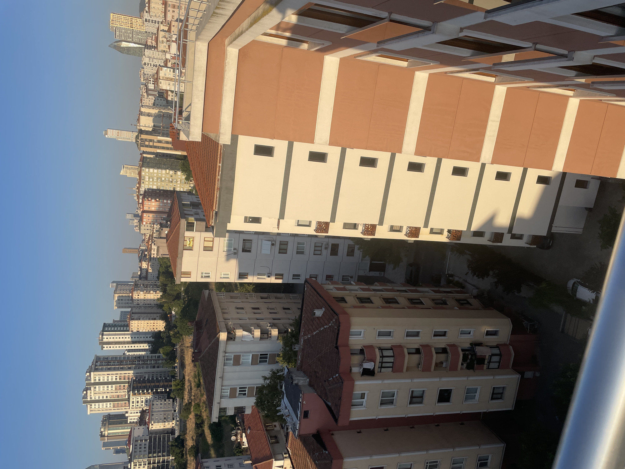 The image captures a cityscape in Kadıköy, Turkey, likely taken from an elevated vantage point. The scene is dominated by residential apartment buildings, varying in height and architectural style. The sky is clear and blue, suggesting a sunny day.
In the foreground, the edge of a metal railing is visible, indicating the viewpoint is from a balcony or window. To the right, a modern apartment building with a terracotta-colored facade and white trim is prominent. Its windows are dark, suggesting they are either shaded or reflecting the sky.
Moving left, a row of multi-story apartment buildings are arranged. Some have lighter, cream-colored facades with horizontal bands of a darker shade and white trim. Others are in shades of yellow and pink with contrasting red details around windows and balconies. These buildings feature numerous balconies, some with ornate railings, and exposed white window frames. Laundry is visible hanging from some balconies.
In the background, a more densely packed urban landscape unfolds. Numerous tall buildings, including some modern skyscrapers with glass facades, rise against the horizon. The architectural diversity suggests a developed urban area. Trees and patches of green vegetation are interspersed between the buildings, particularly on a sloped area in the mid-ground.
The overall impression is of a bustling urban residential area with a mix of older and newer constructions. The lighting suggests it might be morning or late afternoon, with the sun casting long shadows. No people or overt actions are discernible within the scene.