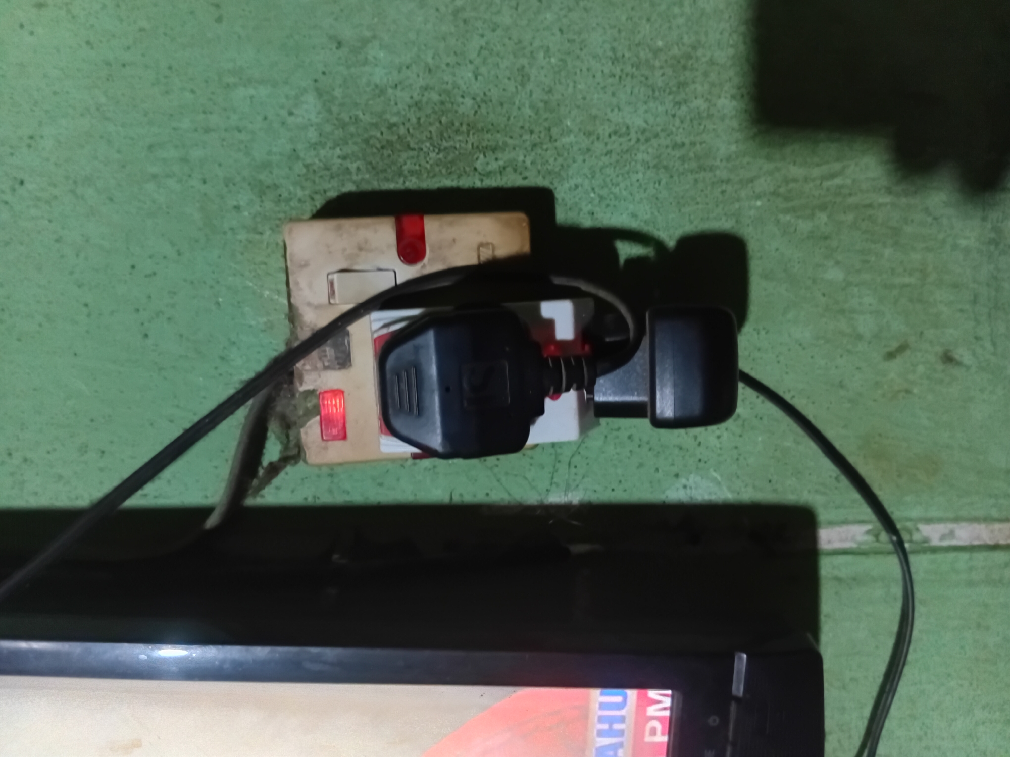 In Nkpor, Nigeria, a close-up shot focuses on a power adapter plugged into a wall socket. The socket, a light beige color, is dirty and mounted on a textured green wall. Two red indicator lights are visible on the socket, one lit and the other off. A black electrical plug is inserted into the socket, connected to a black cable that runs out of frame. A portion of a dark electronic device, possibly a television, is visible on the left side of the image, displaying text that includes "AHU" and "PM". The overall environment appears to be indoors.