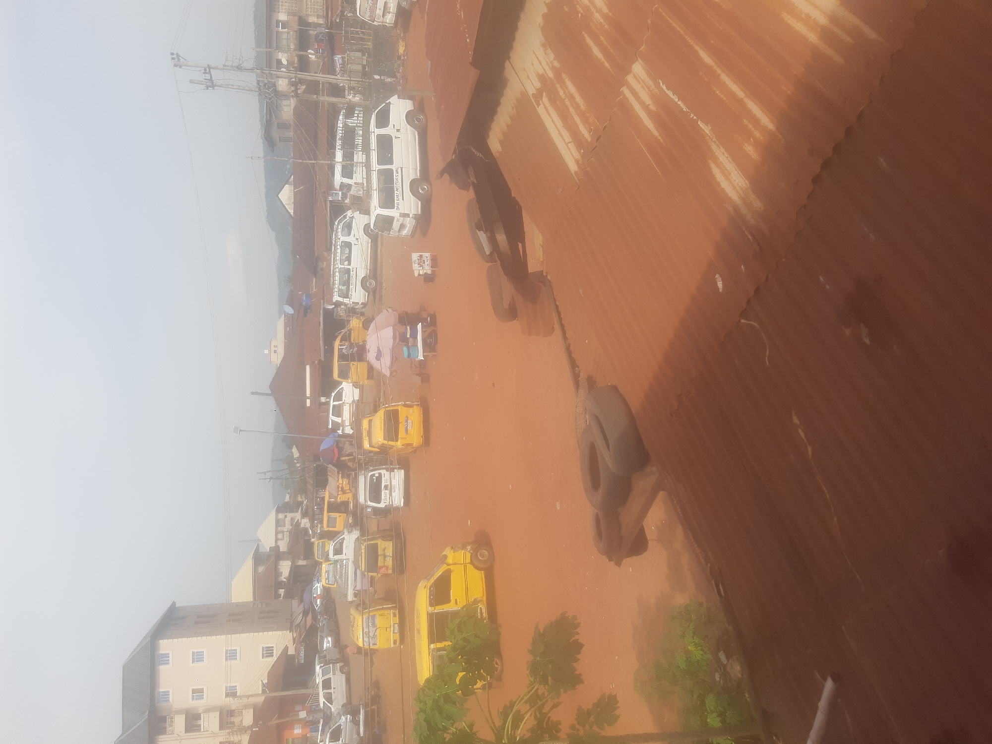 This is a hazy, wide-angle, medium-distance aerial shot of an outdoor scene in a city, likely Enugu, Nigeria, showing a dusty, reddish-brown, unpaved lot filled with parked vehicles. The vehicles are predominantly yellow minibuses, commonly known as "danfo" or "molue" buses, and several white passenger vans. Some vehicles are parked in rows, while others are scattered across the lot. In the background, there are low-rise buildings with corrugated iron roofs and some larger, modern structures. A tall, multi-story building with cream-colored walls and blue-framed windows is visible on the left side of the frame. The sky is overcast and appears hazy, contributing to the overall muted light of the scene. The foreground is dominated by a rusty, corrugated iron roof. A few trees, including one with large green leaves on the left, are present. There are no discernible individuals performing actions in the image, and the overall impression is of a busy transport hub or parking area in a developing urban environment.