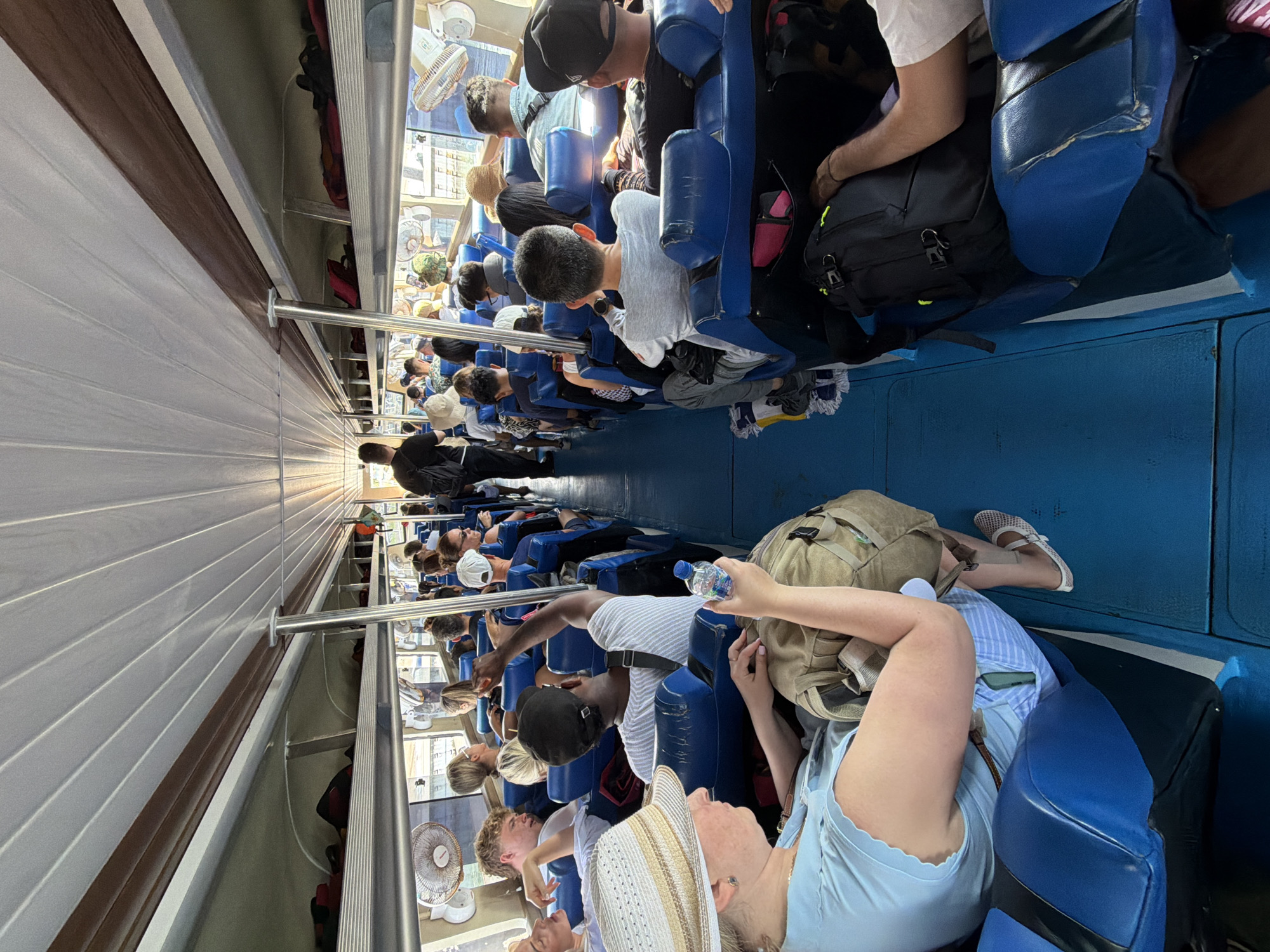 The image depicts the interior of a crowded ferry or bus, likely in Denpasar, Indonesia, with rows of blue seats occupied by passengers. The ceiling is made of white, ribbed panels with brown trim. On the right side, a man in a black t-shirt and a backpack is walking down the aisle. On the left, a woman wearing a straw hat and a light blue top is seated, holding a water bottle and a backpack. Other passengers, varying in age and ethnicity, are visible throughout the vehicle, many with luggage. Small fans are mounted on the walls near the windows. The overall impression is one of a busy journey.
