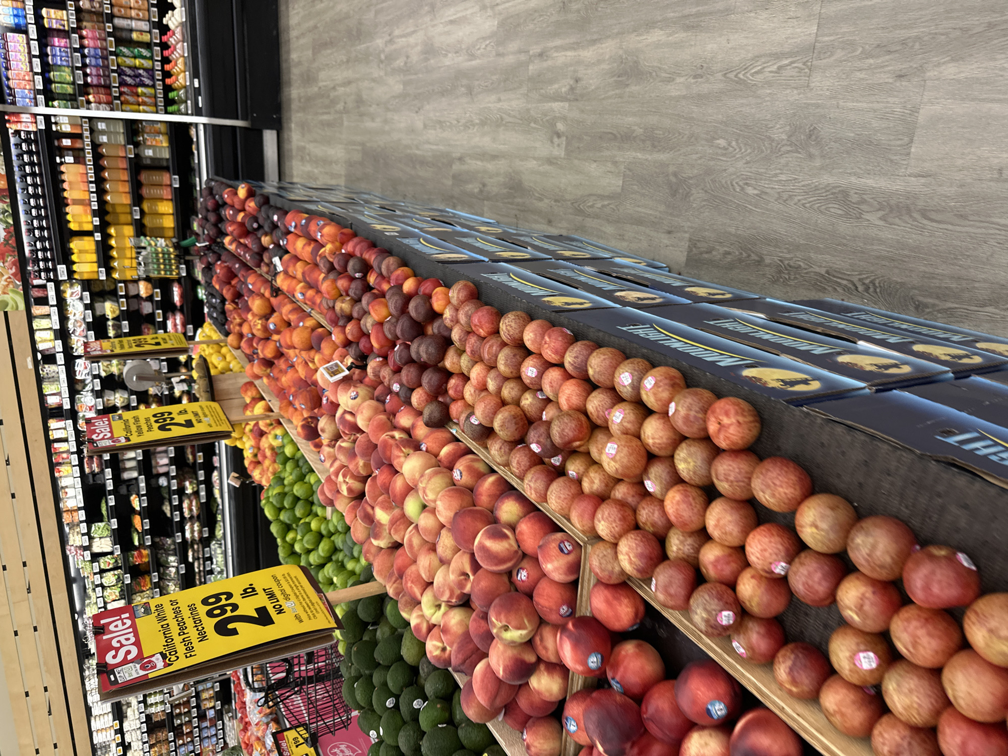 The image shows a produce aisle in a grocery store, likely a Mariano's or a similar supermarket, located in Schaumburg, United States of America. The aisle is filled with various fruits and vegetables displayed on tiered shelving and bins. Prominently featured are large bins of avocados, limes, and several varieties of peaches and nectarines. Various signage indicates sales, with one sign prominently displaying "California White Flesh Peaches or Nectarines" for $2.99 per pound. The background reveals shelves stocked with bottled beverages, and refrigeration units further down the aisle. The flooring is a light grey wood-grain laminate. No people are visible in the scene, and the focus is entirely on the retail display of produce.