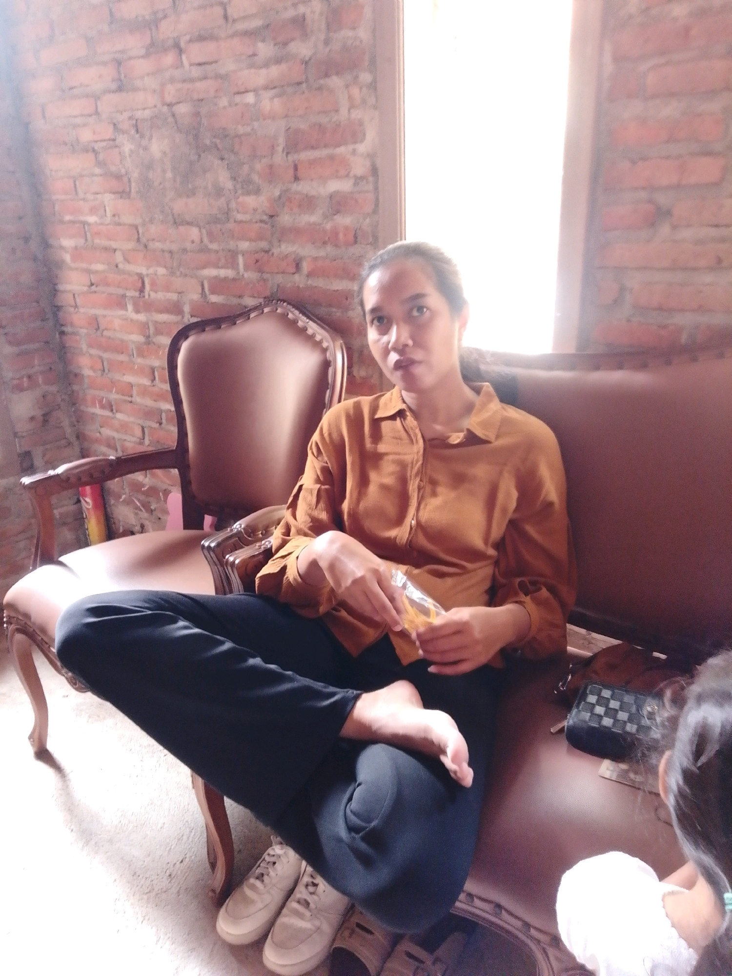 An adult, dressed in a rust-colored button-down shirt and black pants, sits cross-legged on a brown sofa in a room with a brick wall. The person is holding a clear plastic packet with yellow contents. A second person, partially visible from behind, sits to the right on the sofa, wearing white. A black checkered clutch bag is on the sofa next to the first person. A wooden chair with brown upholstery is to the left of the sofa. The room is illuminated by light from a window on the right side of the image. The location is identified as Cikal, Indonesia.