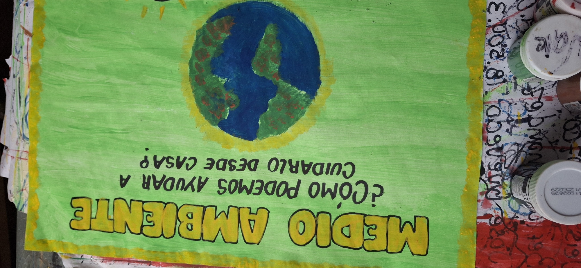The image is a close-up shot of a handmade poster about environmental awareness. The poster is primarily green with a yellow border. A stylized image of the Earth, with blue oceans and green landmasses outlined in yellow, is in the upper section. Text in Spanish reads "MEDIO AMBIENTE ¿CÓMO PODEMOS AYUDAR A CUIDARLO DESDE CASA?" (ENVIRONMENT. HOW CAN WE HELP CARE FOR IT FROM HOME?).
Located to the right of the poster are several paint containers with white lids, some of which have paint stains or writing on them. The surface underneath appears to be covered in paint, numbers and text.
There is no indication that the photograph was taken in Vallecito, Venezuela. There are no people present in the image.